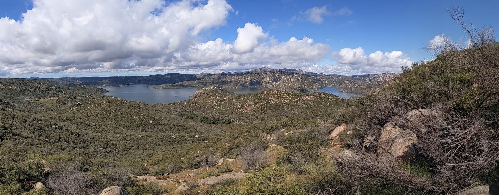 Panoramic view of a lake and rocky hills under a bright, partly cloudy sky.