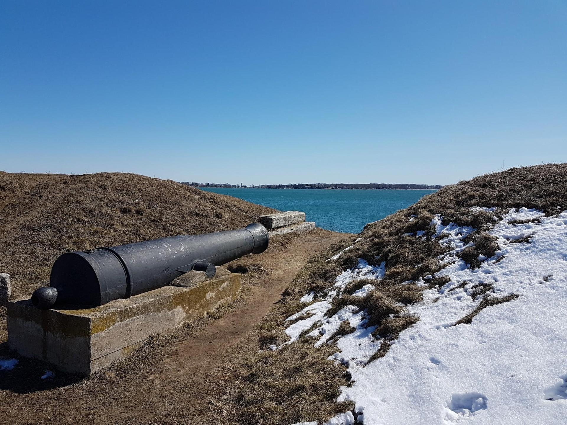An old cannon on a snowy, grassy hill overlooking blue water.