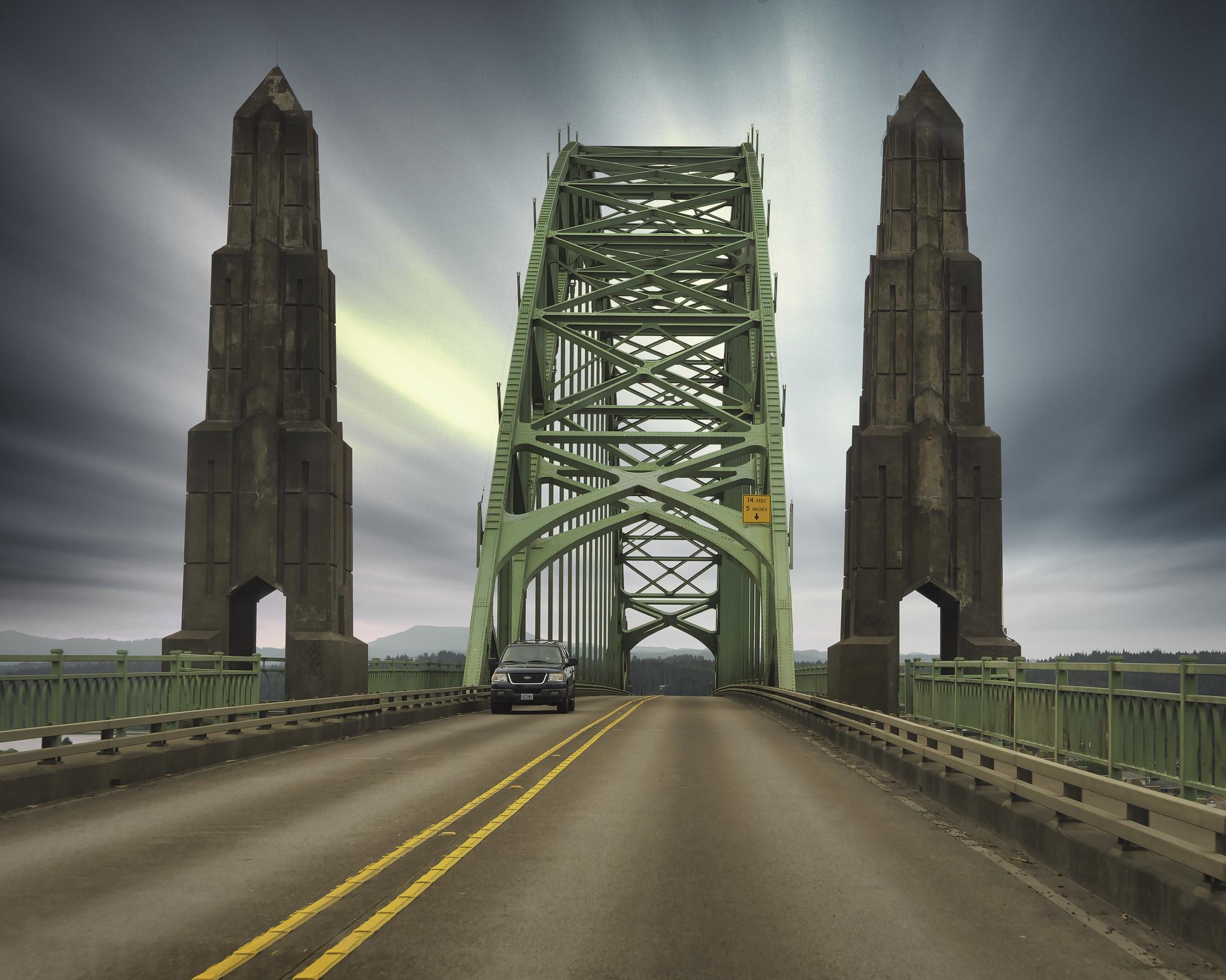 Dramatic view of a green steel bridge with tall concrete towers and a car.