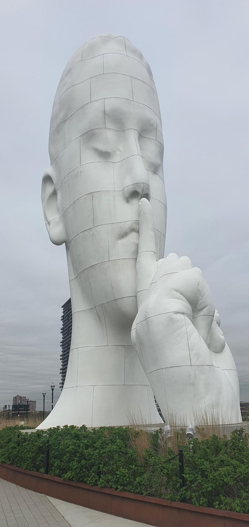 Giant white head sculpture with a finger to its lips, under an overcast sky.