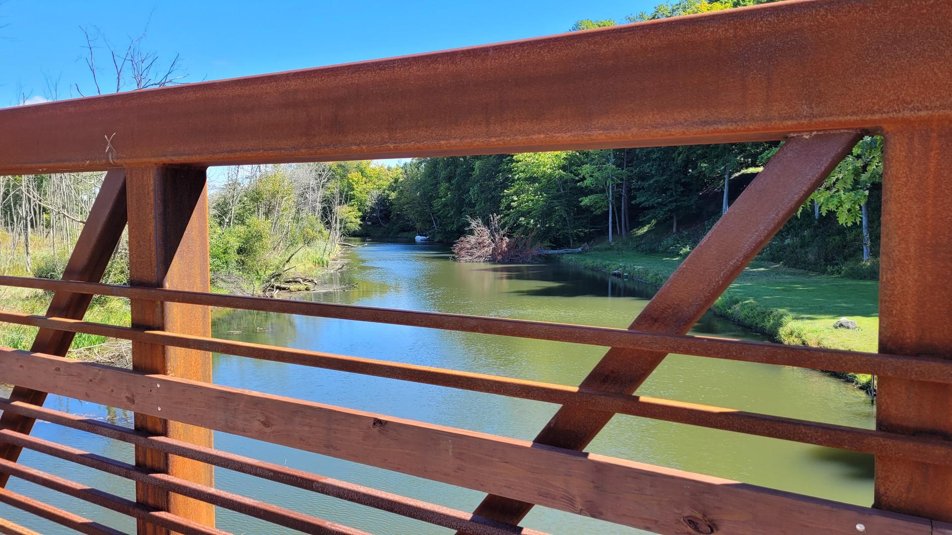 Sunny view of a calm river through a rusty metal bridge railing.