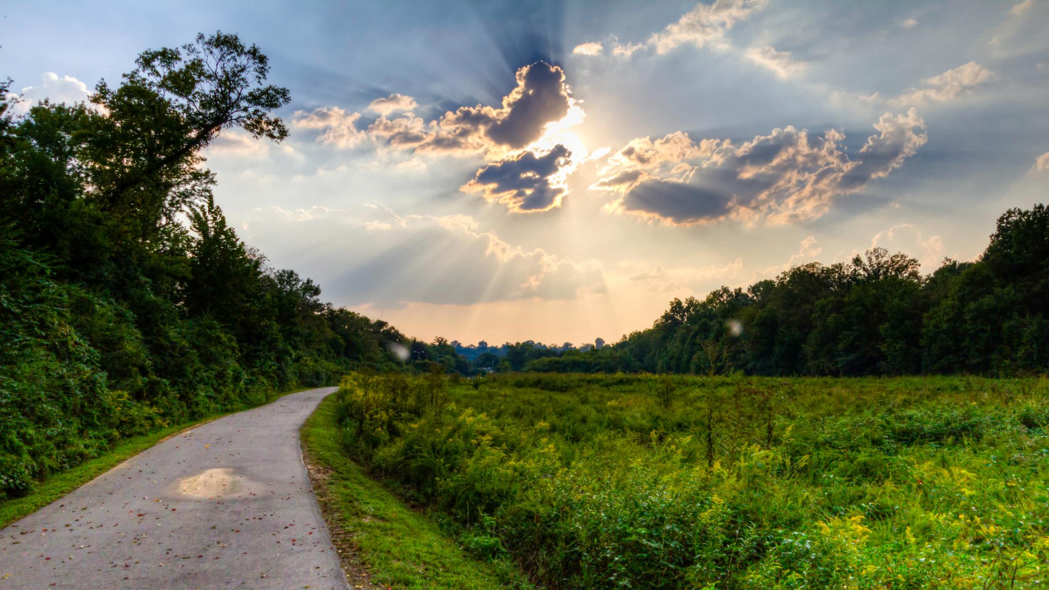 Paved path curves through a sunny green field with sun rays breaking through clouds.
