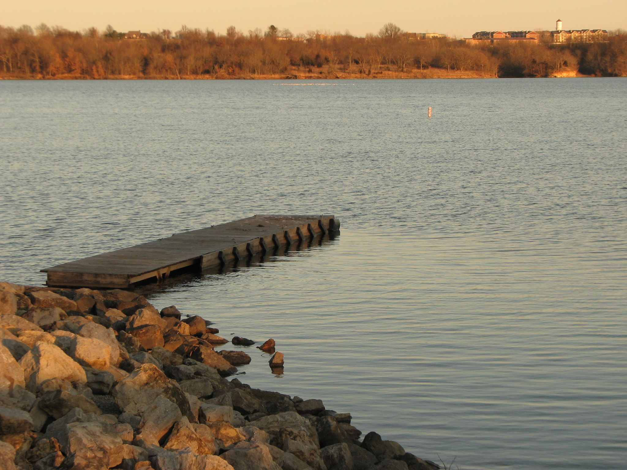 A wooden dock extends into calm water with a rocky shore at sunset. - Longview Lake Trail