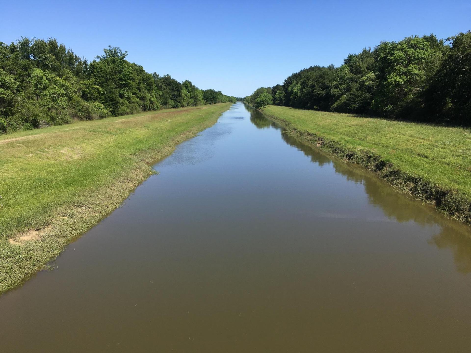 A long, straight canal with grassy banks and green trees under a clear sky.