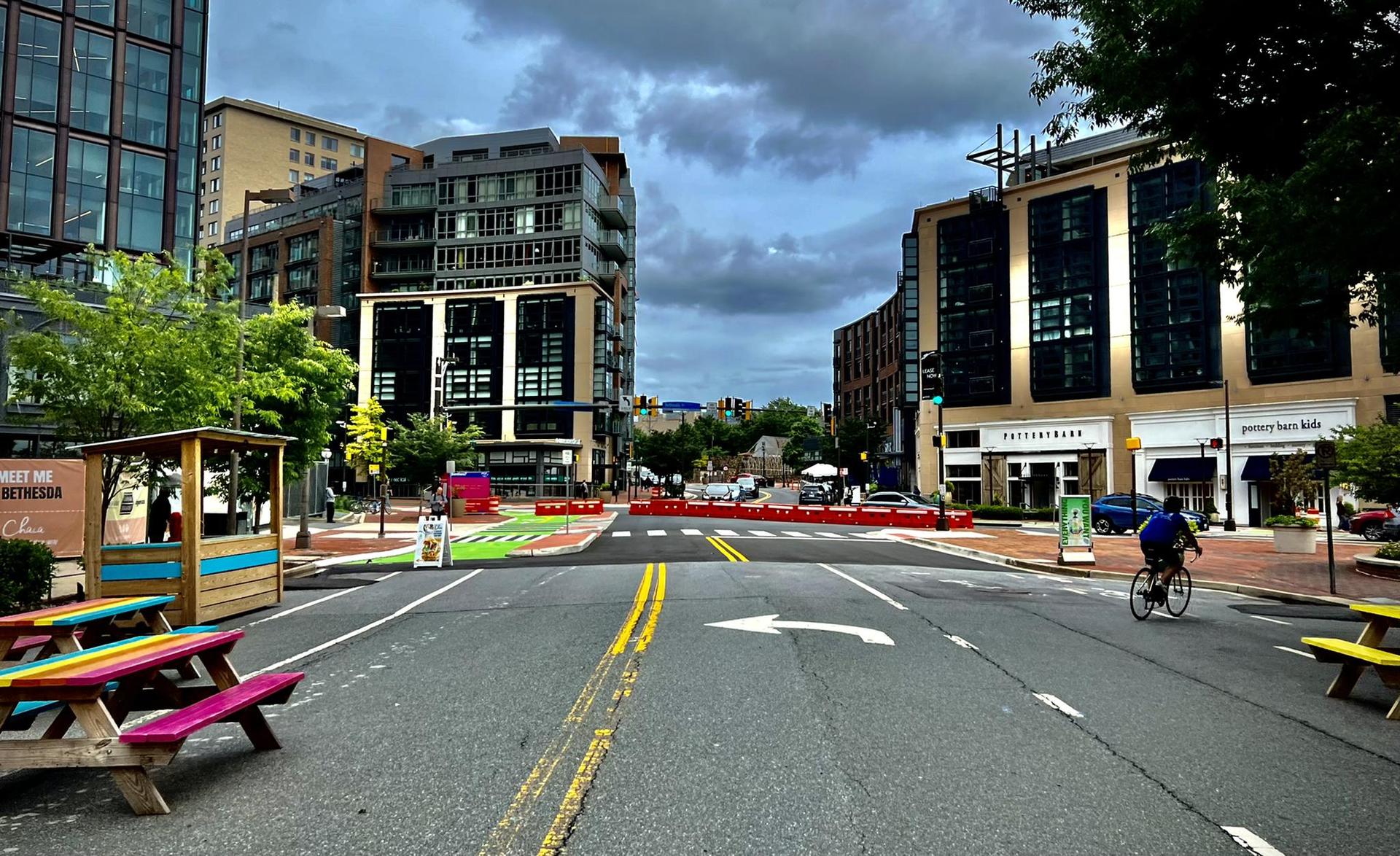 City street with modern buildings, colorful picnic tables, and a cyclist.