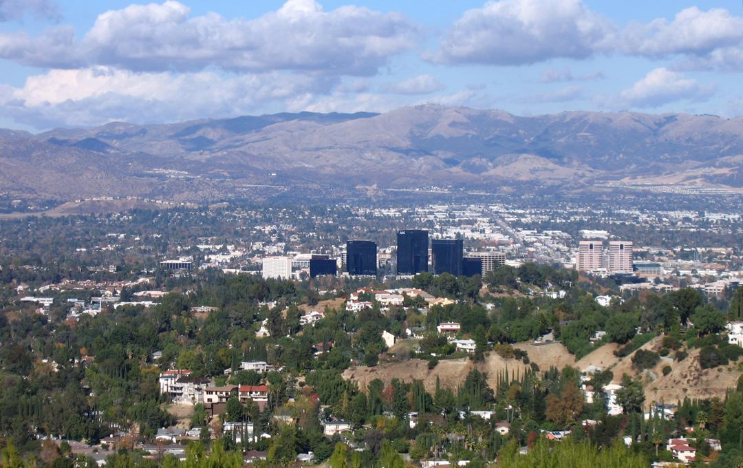 Expansive view of a city nestled among green hills and distant mountains.