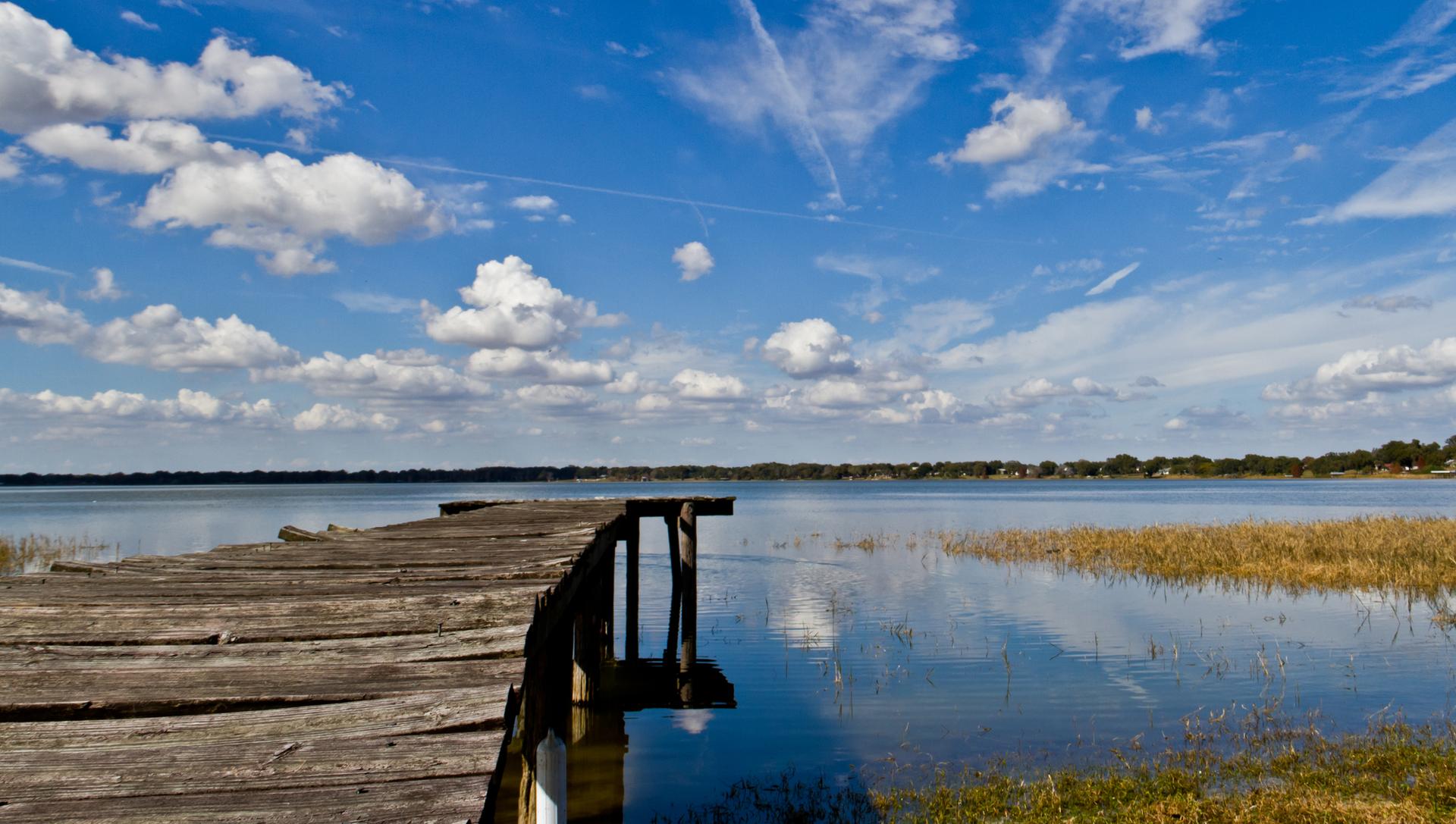 A weathered wooden dock extends into a calm lake under a bright blue sky.