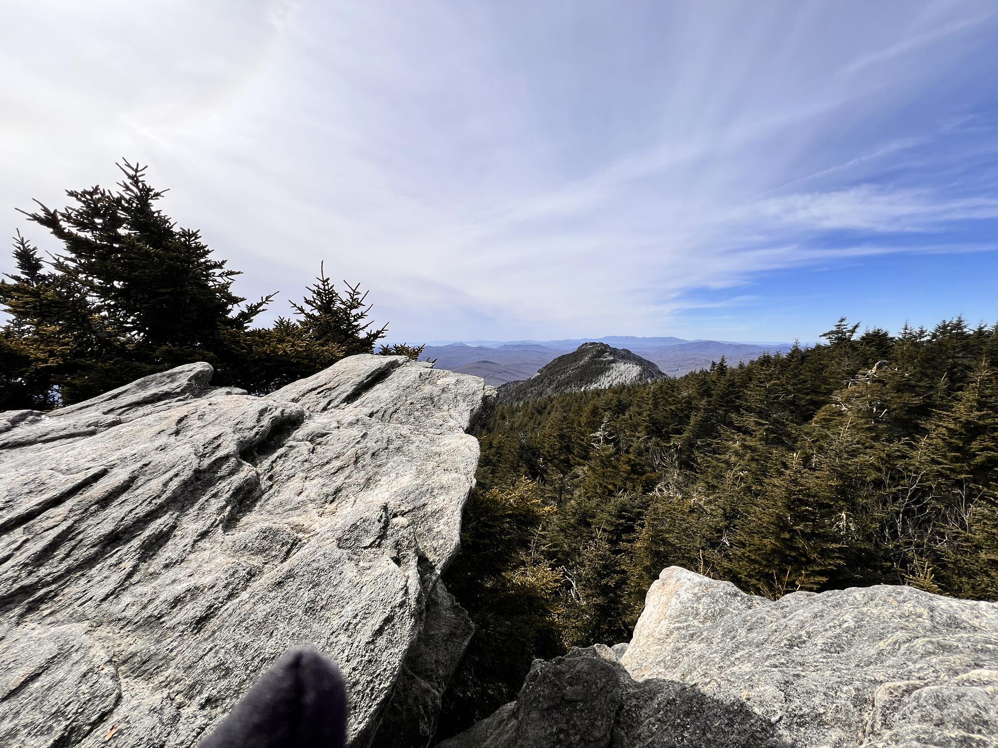 Person's foot on rocky outcrop overlooking a vast forest and distant mountains.