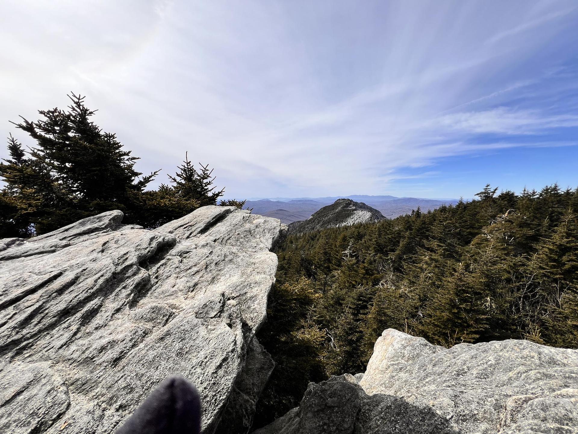 Person's foot on rocky outcrop overlooking a vast forest and distant mountains.