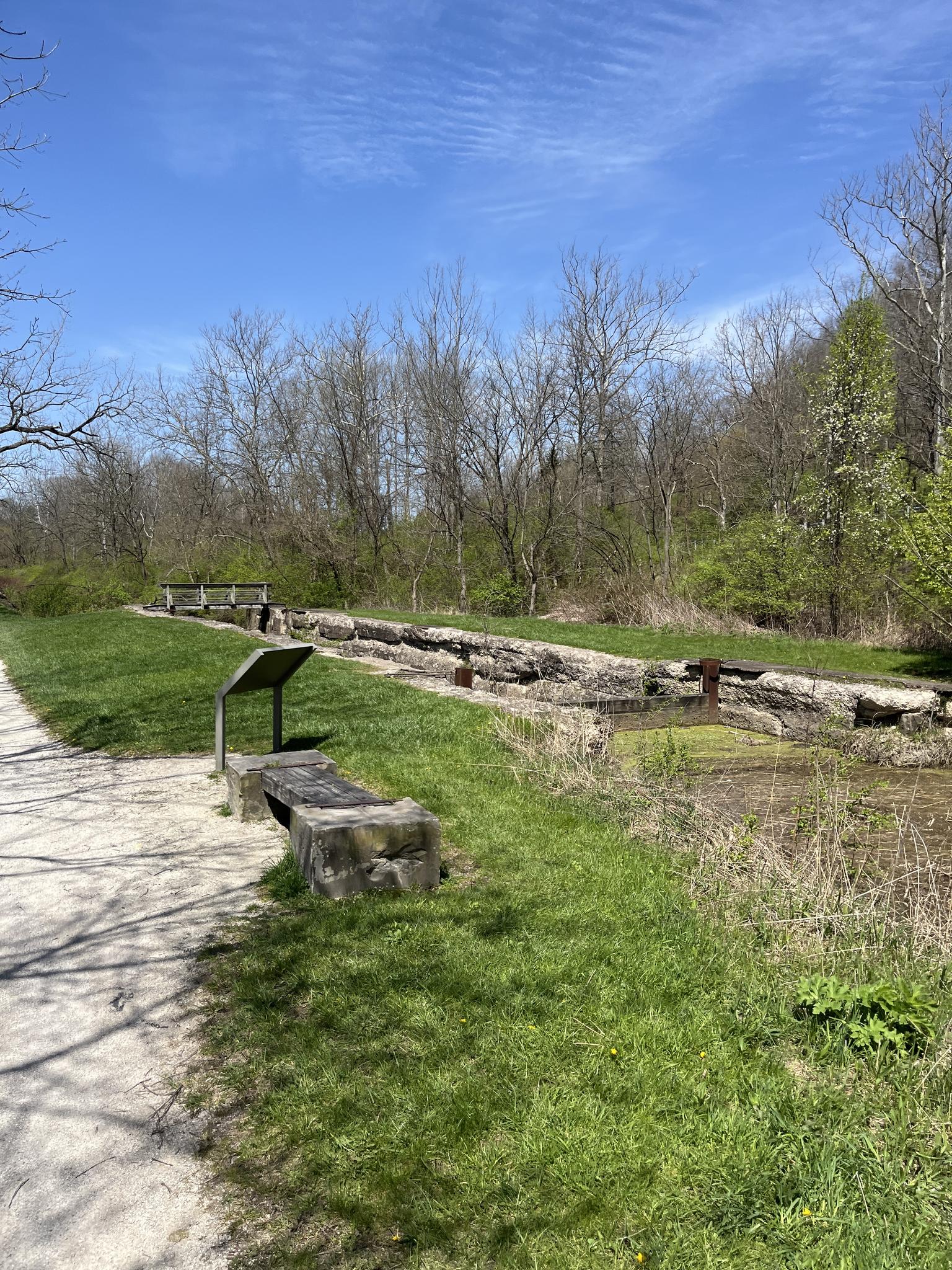 Long, overgrown canal lock structure next to a path under a blue sky.
