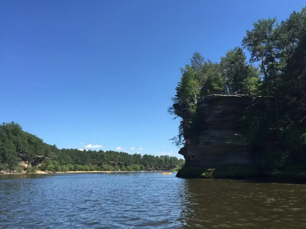 Sunny river with a rocky cliff, dense trees, and distant kayaks.