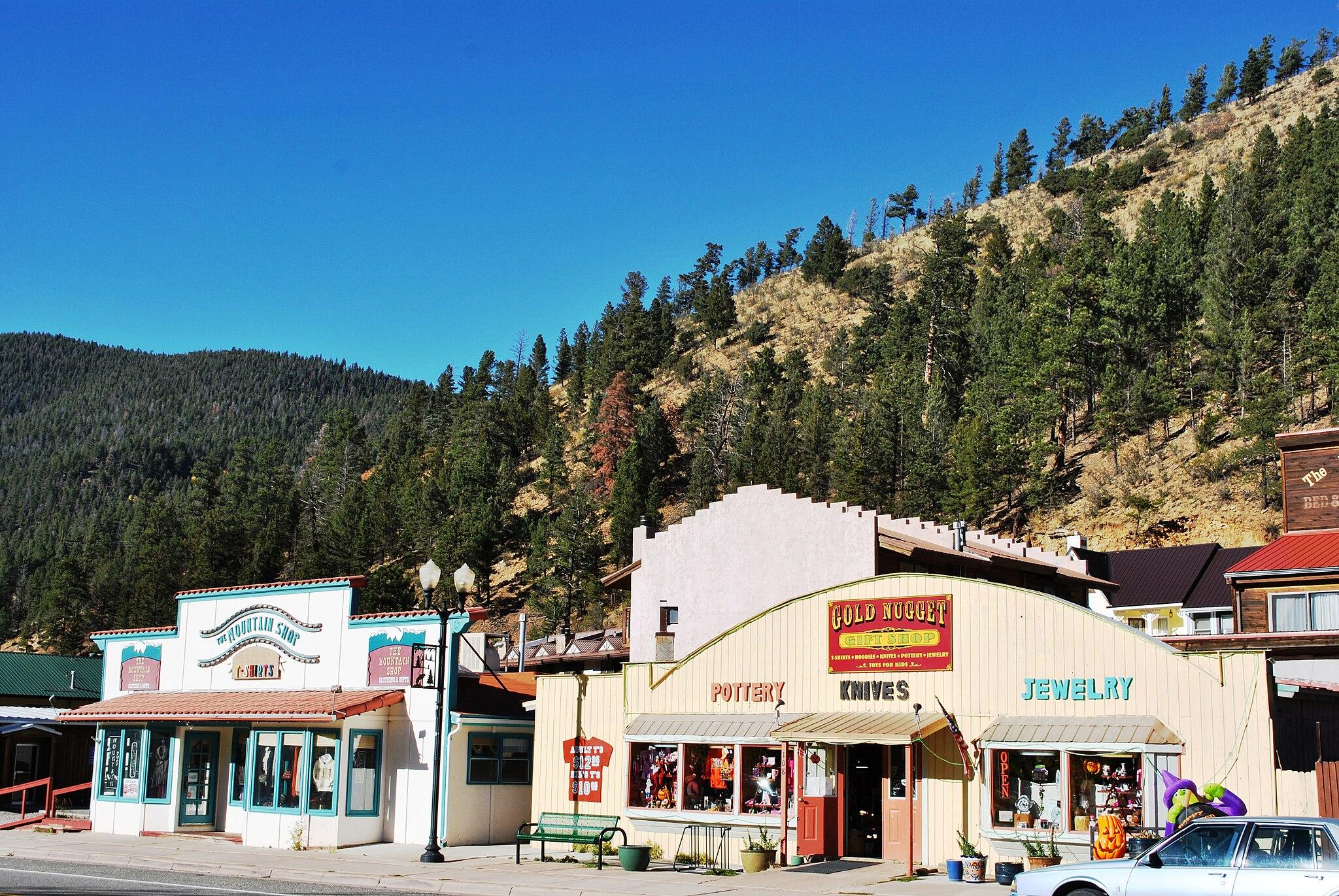 Small town buildings line a street with a steep, pine-covered mountain under bright sun.