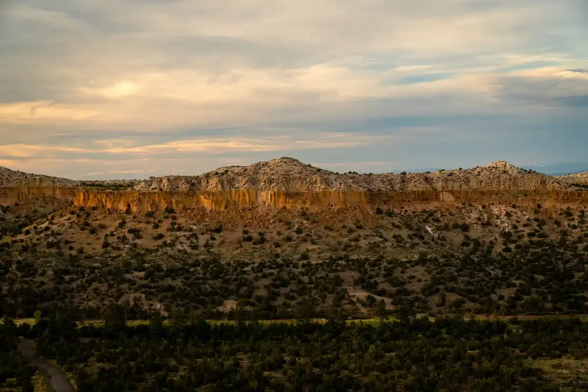Golden light illuminates rocky desert cliffs and hills under a cloudy sky.