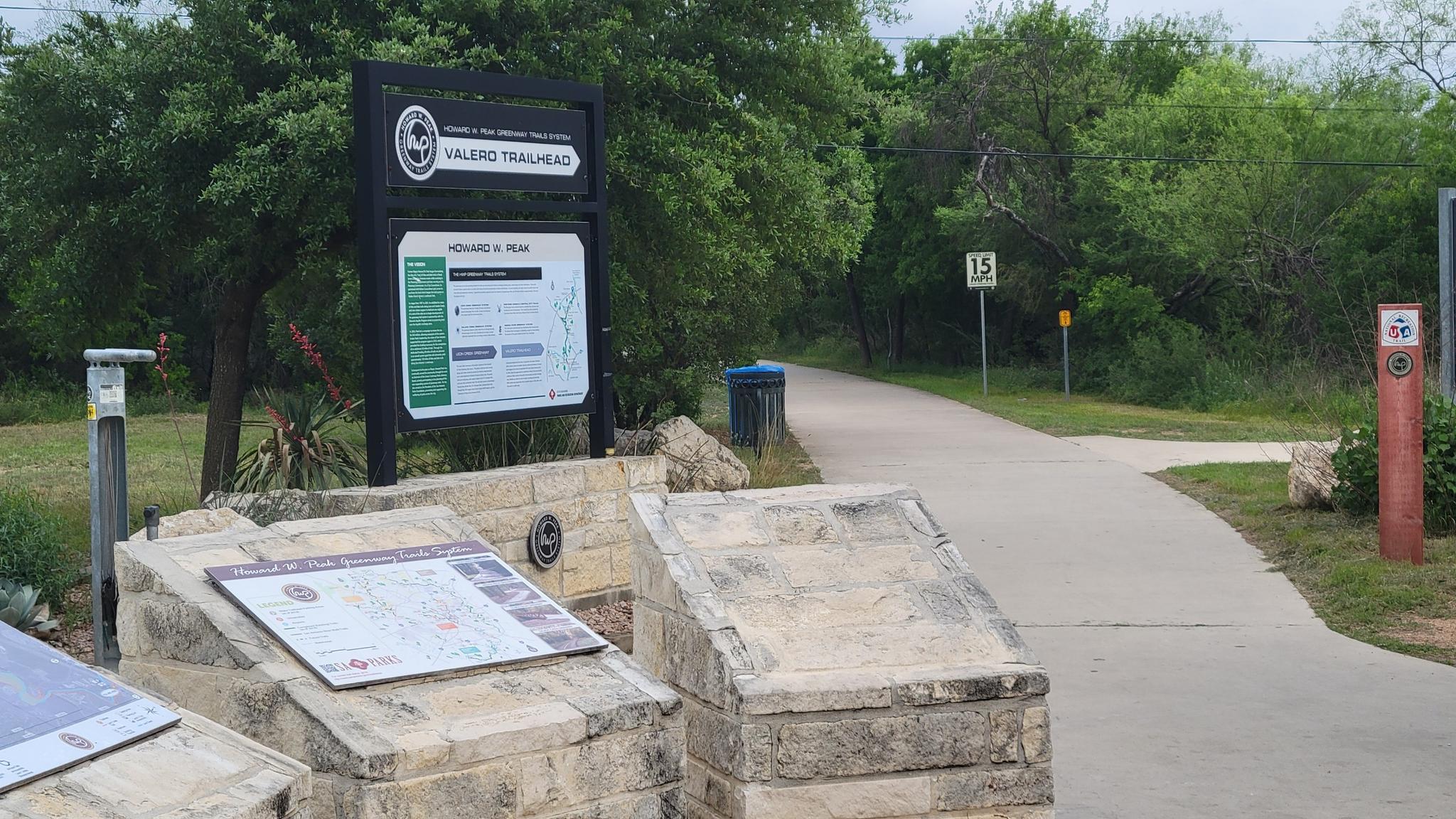 Trailhead sign and information boards next to a paved path on an overcast day.