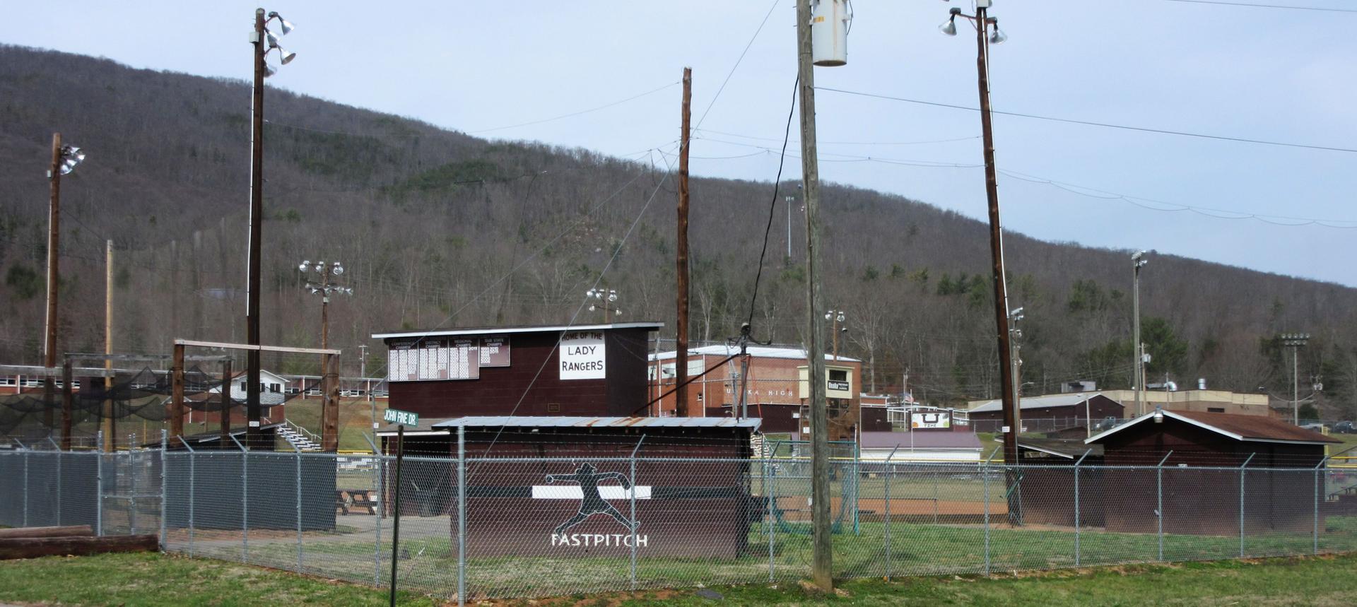 An empty baseball field with dugouts and light poles, backed by a wooded mountain.