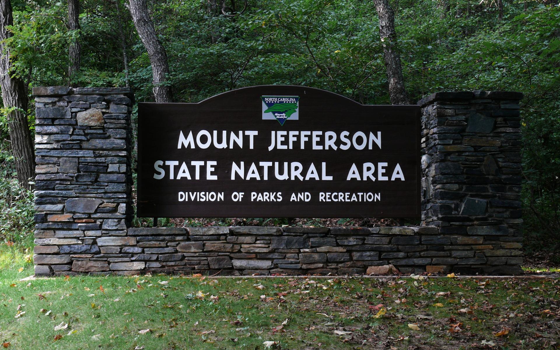 A large dark sign with white lettering on stone pillars, surrounded by green forest.