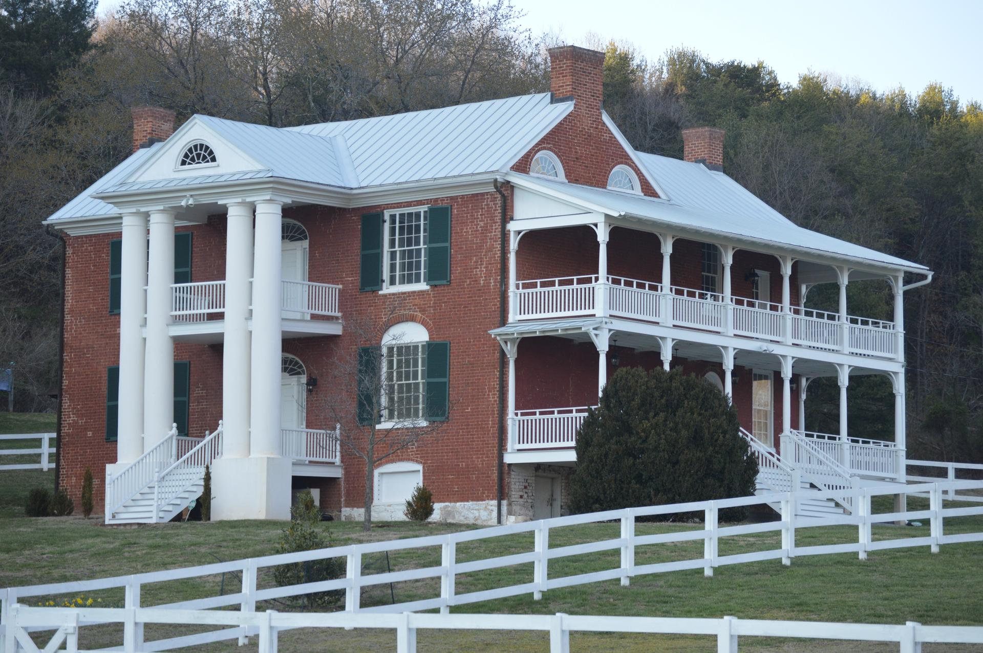 A large, historic brick house with white columns and porches on a grassy hill.