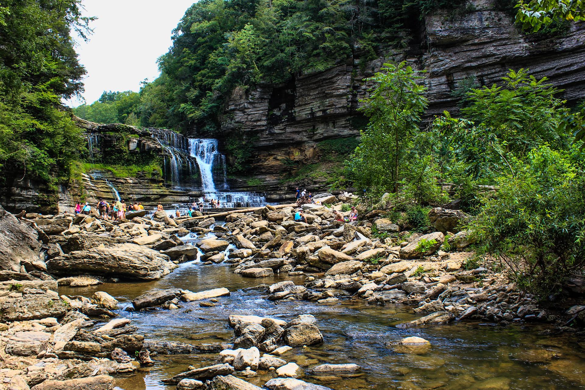 People exploring a rocky riverbed below a waterfall in a green gorge.