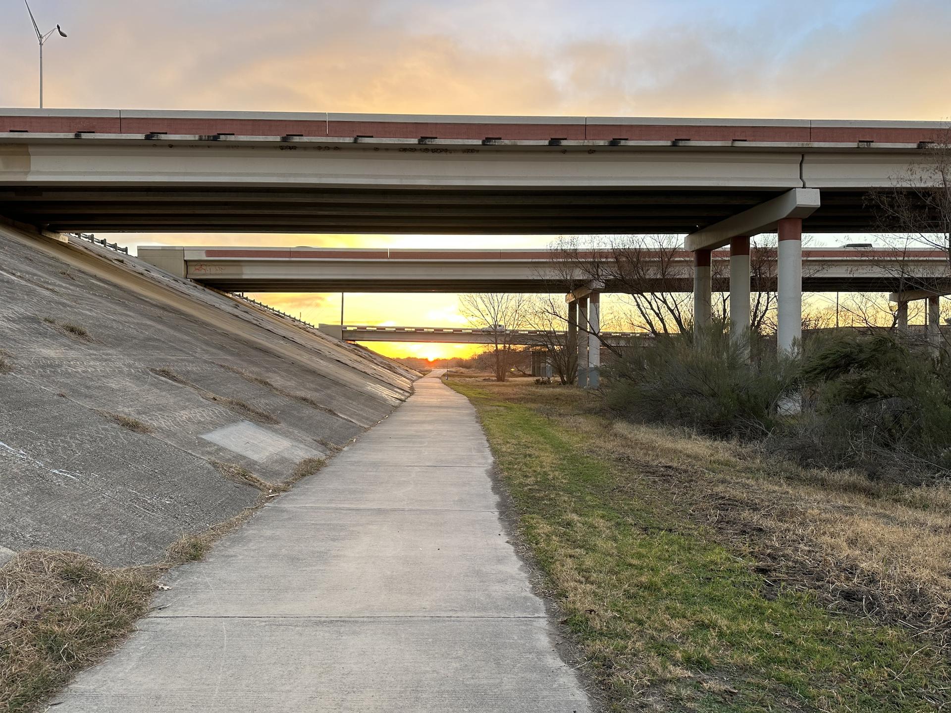 A paved path extends under highway overpasses at sunset.