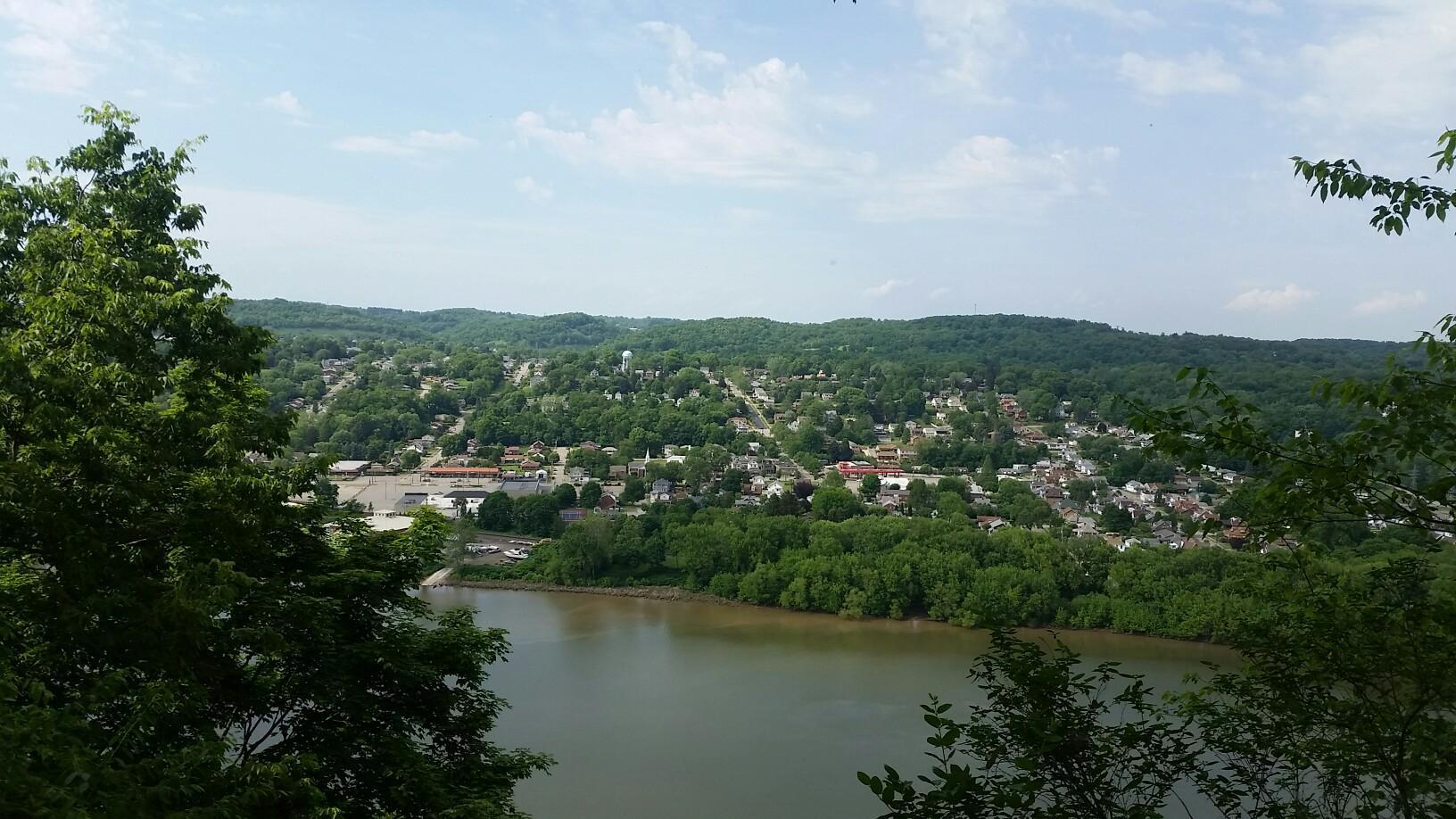 Panoramic view of a river, town, and forested hills under a bright sky.