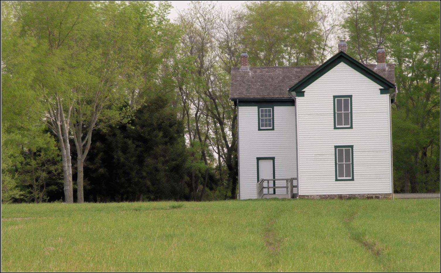 White two-story house with green trim in a grassy field, surrounded by trees.