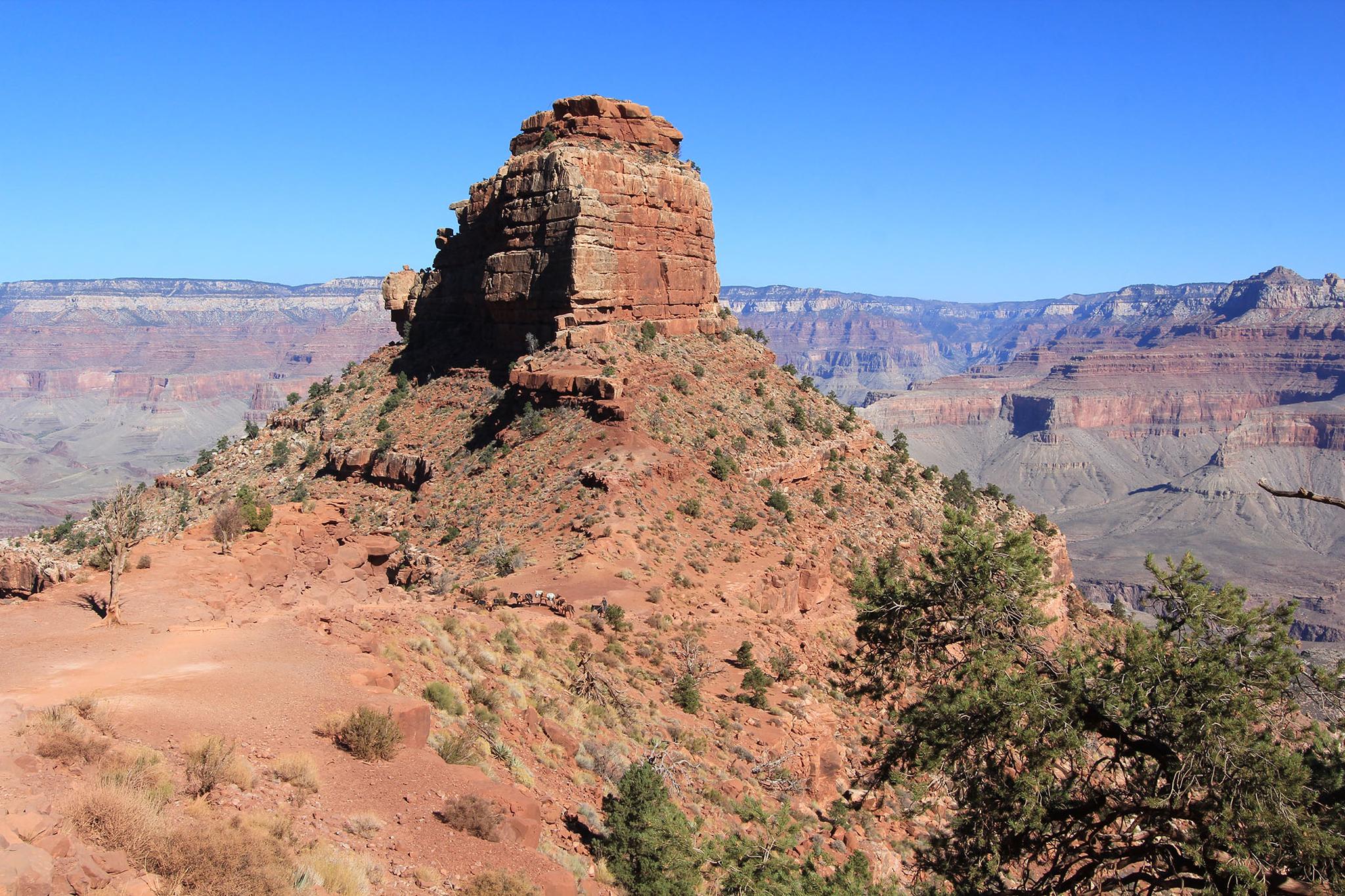 Large, red rock formation dominates an arid canyon landscape under blue sky.