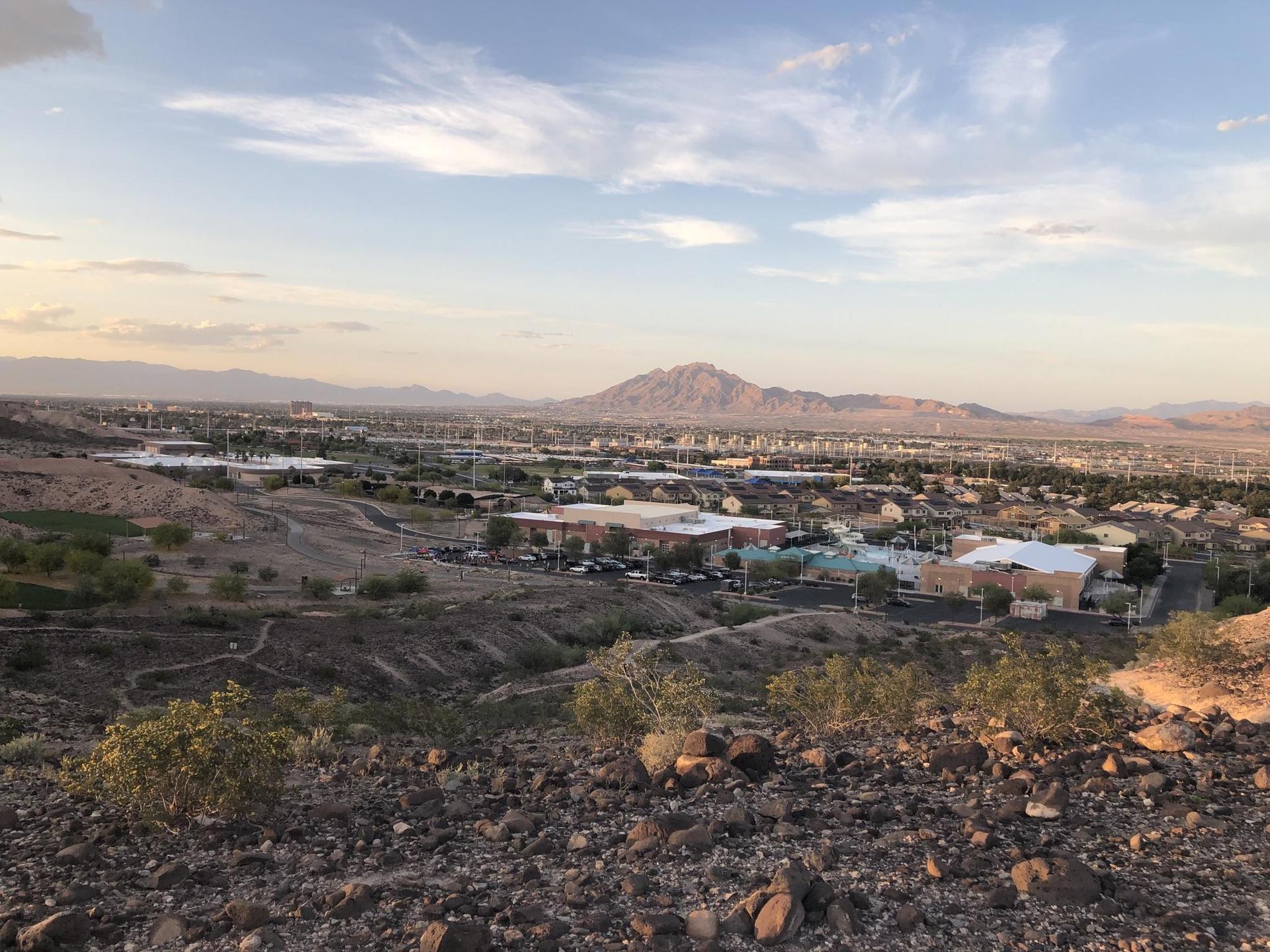 Rocky desert foreground overlooks a town with distant mountains under a cloudy sky.