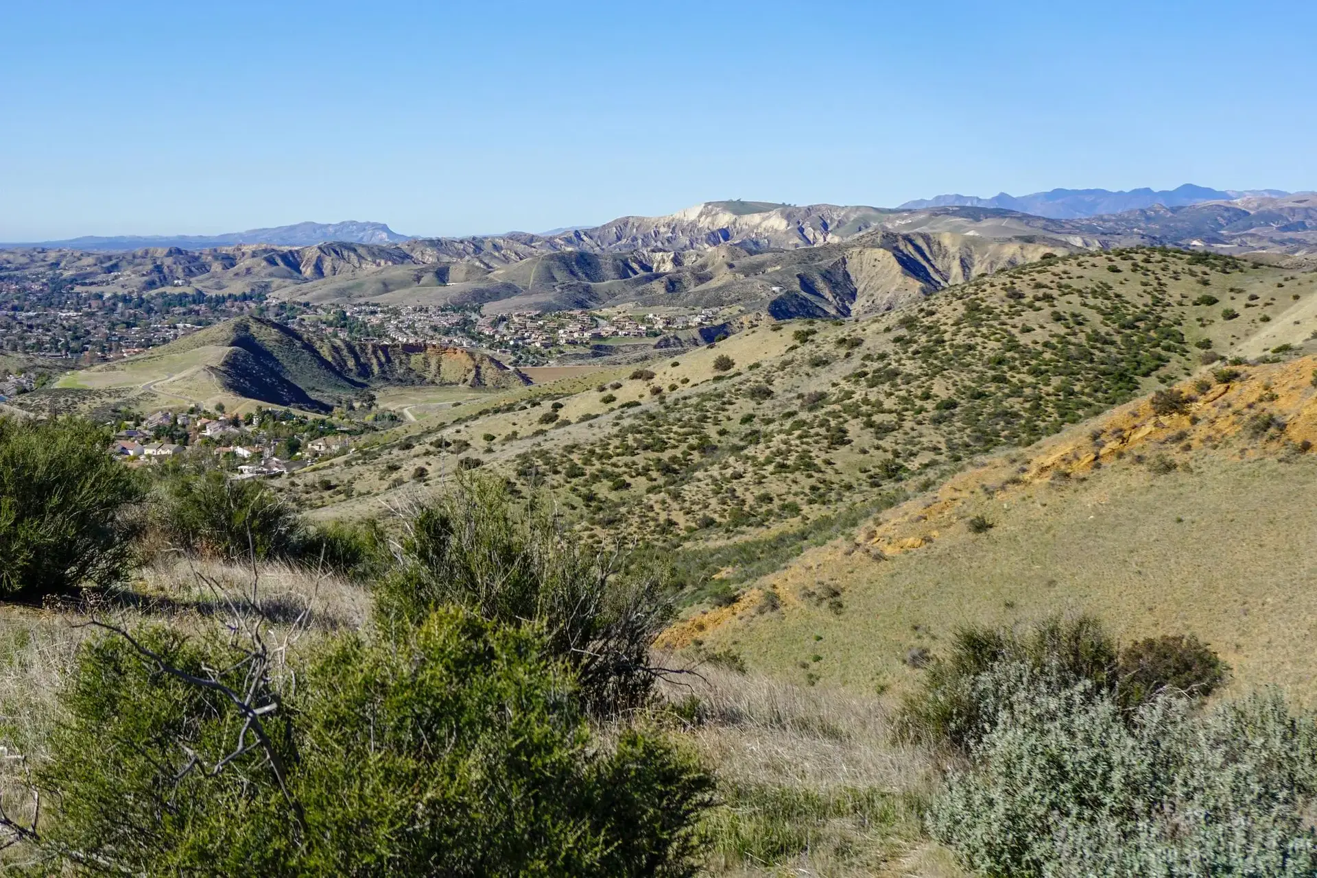 Expansive view of sunny, dry rolling hills with scattered green brush and distant buildings.