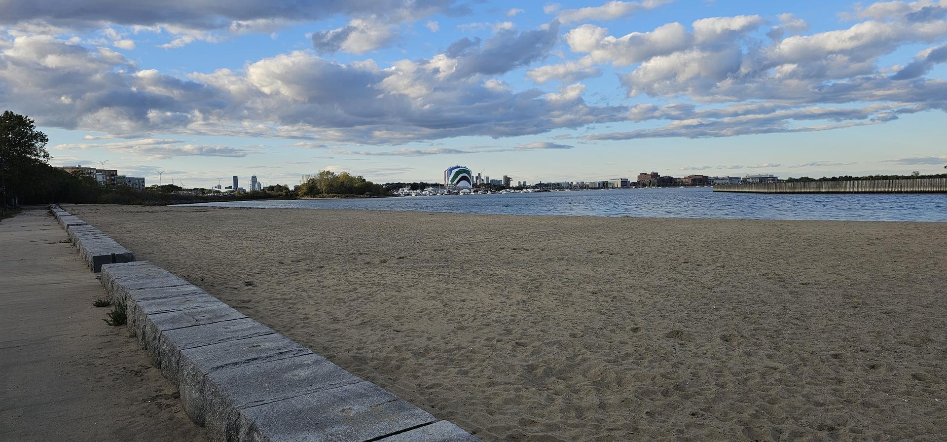 A wide, empty sandy beach next to calm water under a cloudy sky.