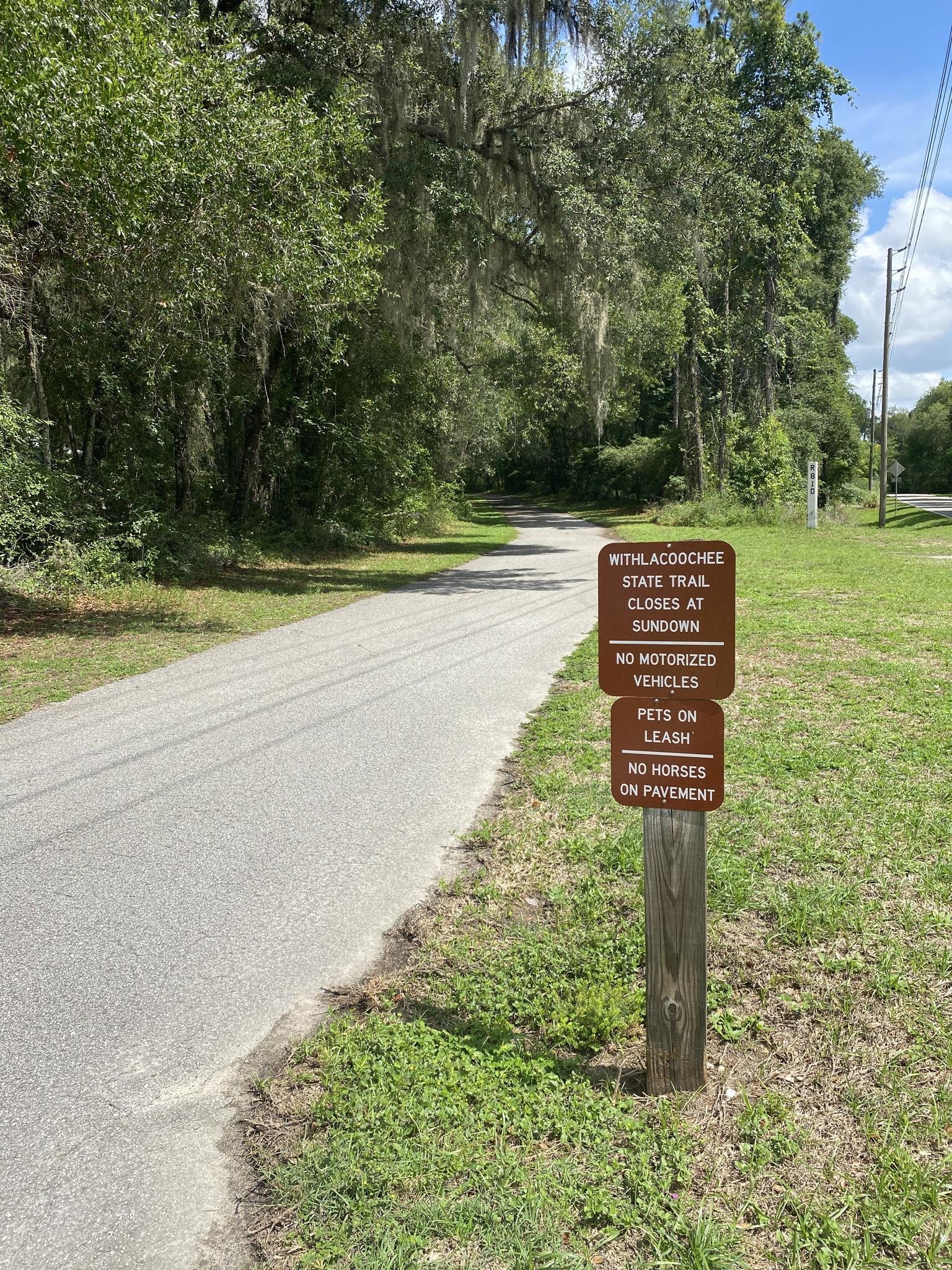 Paved path next to a brown sign with rules, surrounded by trees.