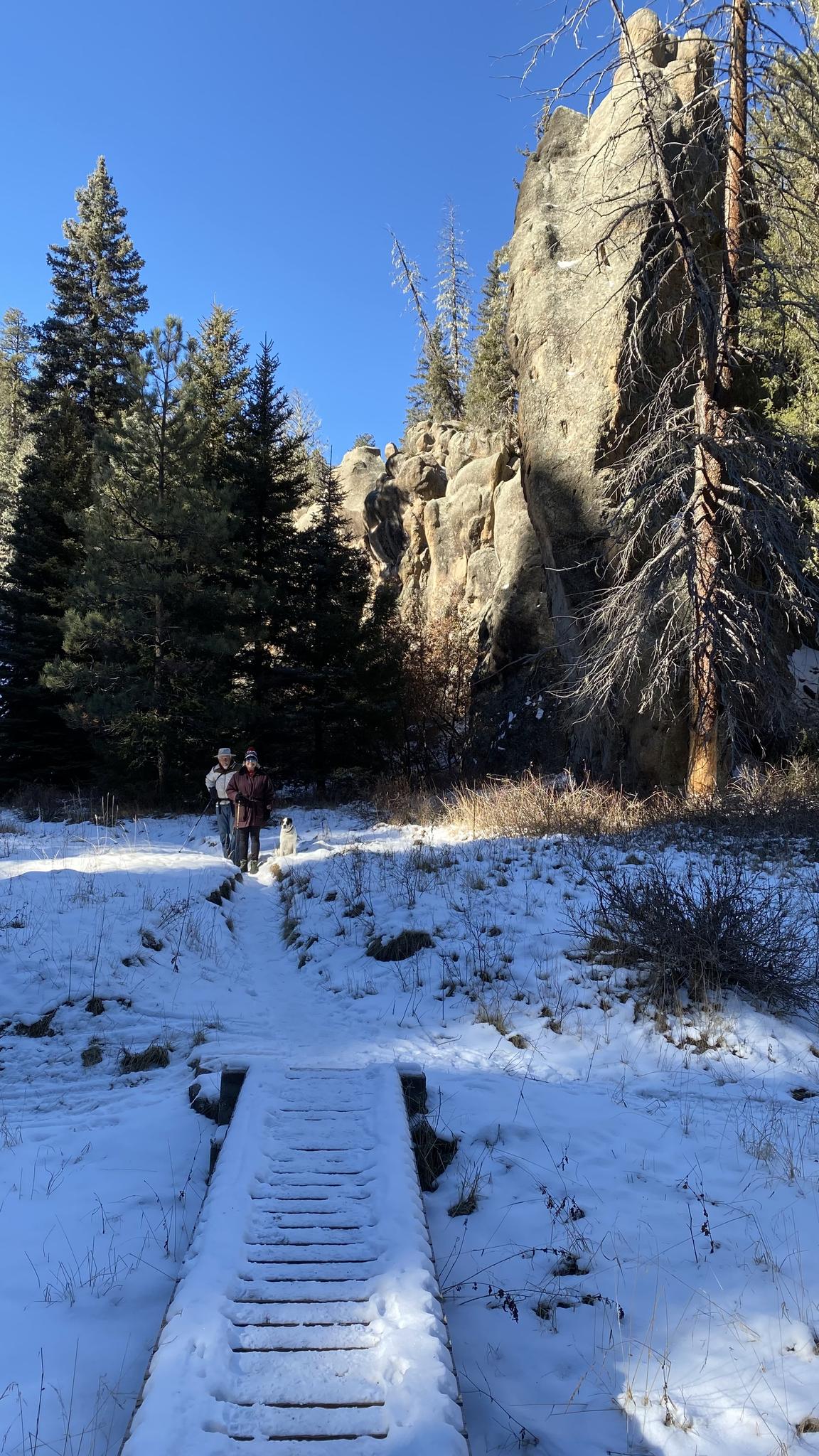 Two people hike on a snowy path leading to a wooden bridge and rock formations.