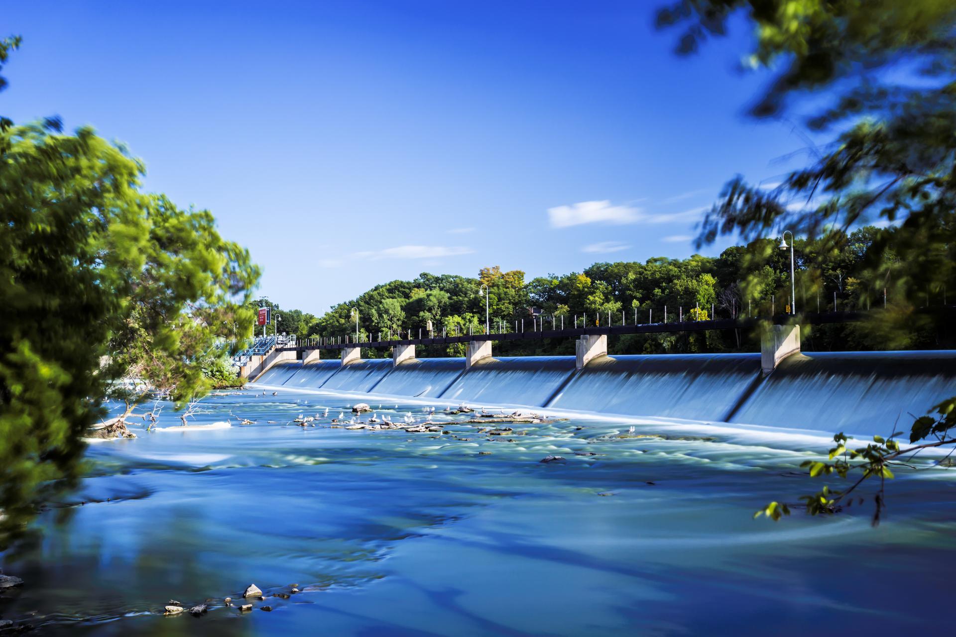 Wide dam with water flowing over it, lush green trees, bright blue sky.