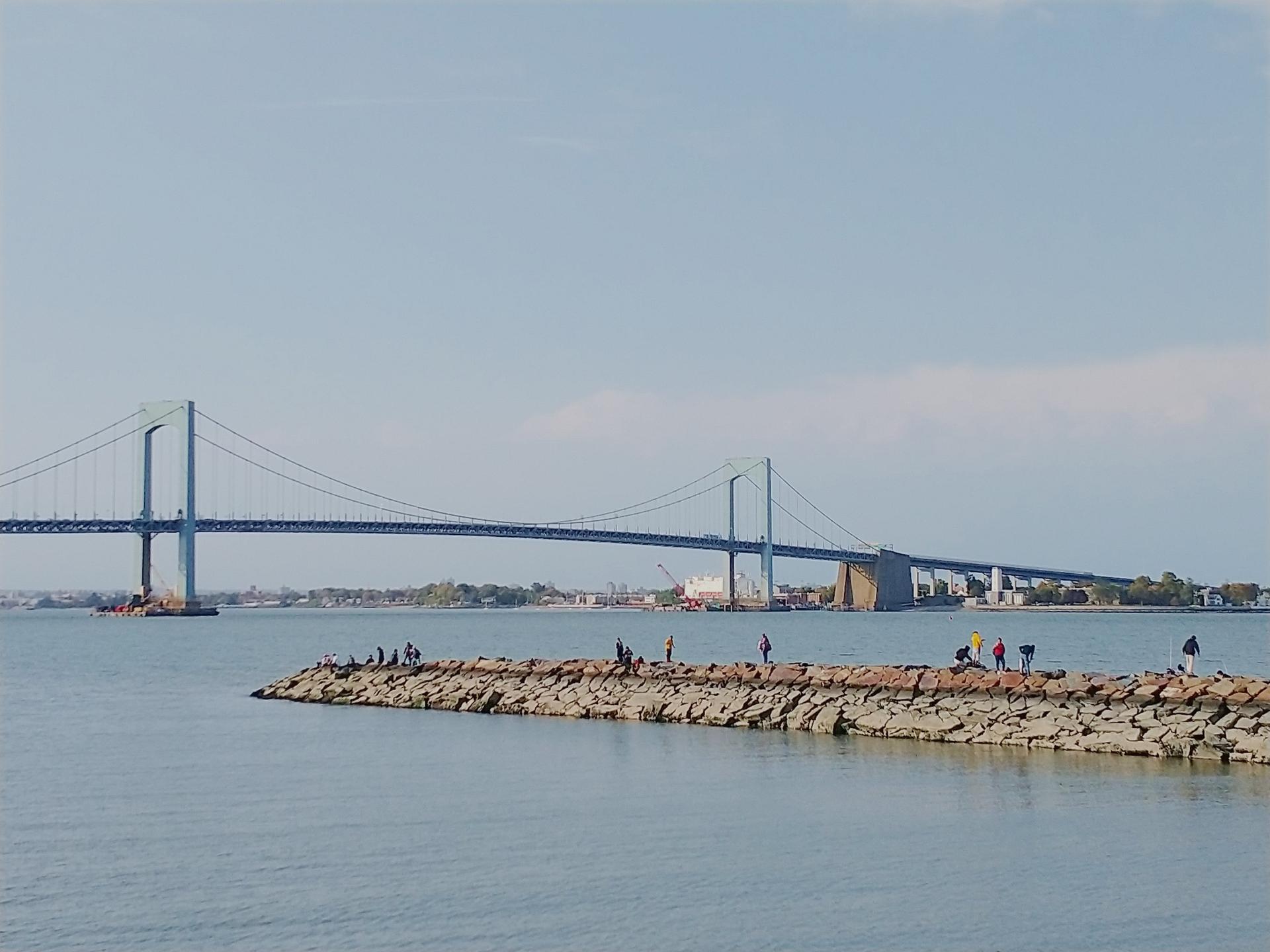 Numerous people on a rocky jetty with a large suspension bridge in the distance.