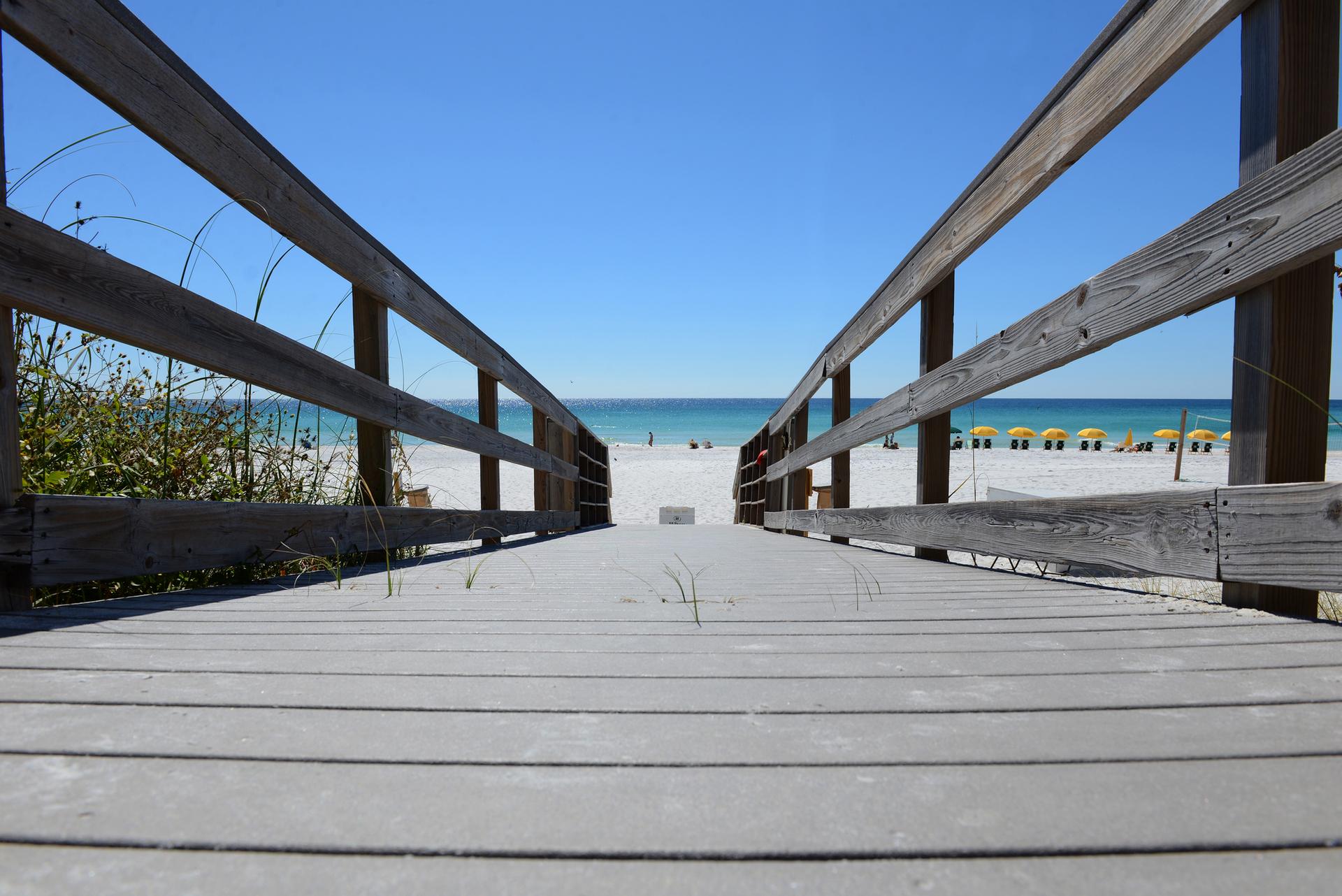 Wooden boardwalk leading to a sunny white sand beach with distant people.
