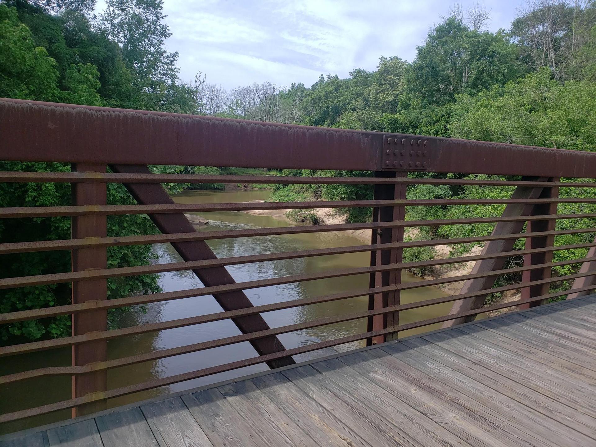 View from a wooden bridge deck looking through metal railings at a muddy river.