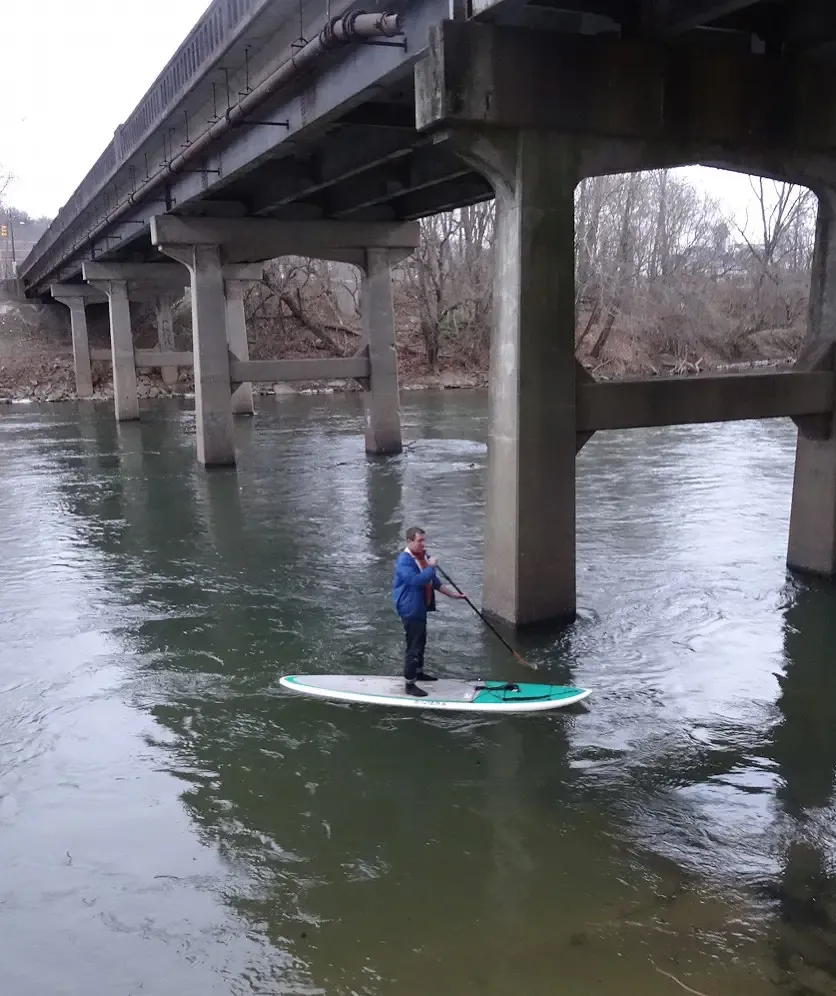Person paddleboarding on a river under a large concrete bridge. - Jean Webb Park – Carrier Park loop from Silver Line Station