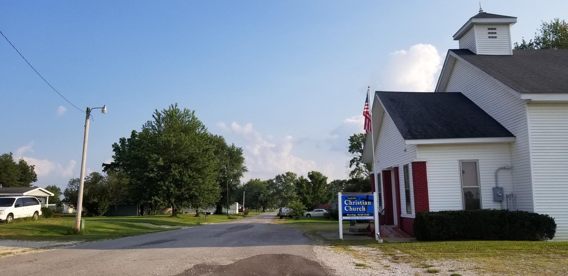 White church building and street under bright blue sky with scattered clouds.