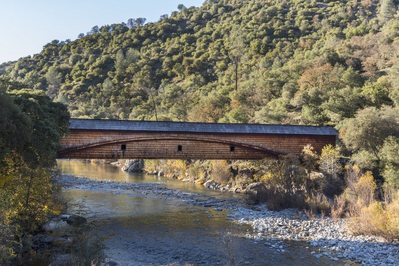 A wooden covered bridge spans a rocky river with forested hillsides under daylight. - Buttermilk Bend Trail