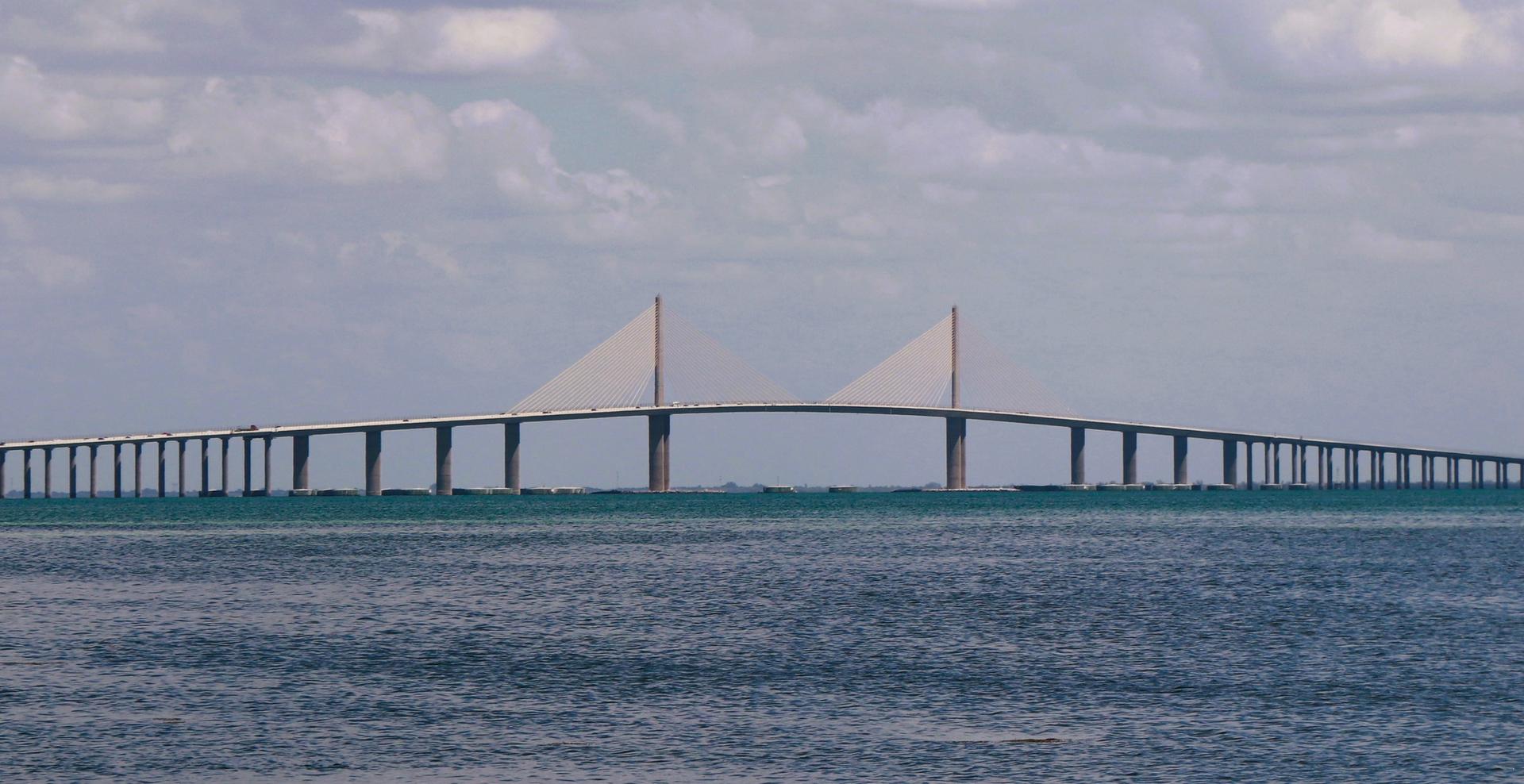 Long cable-stayed bridge spans across a wide body of water under a cloudy sky.