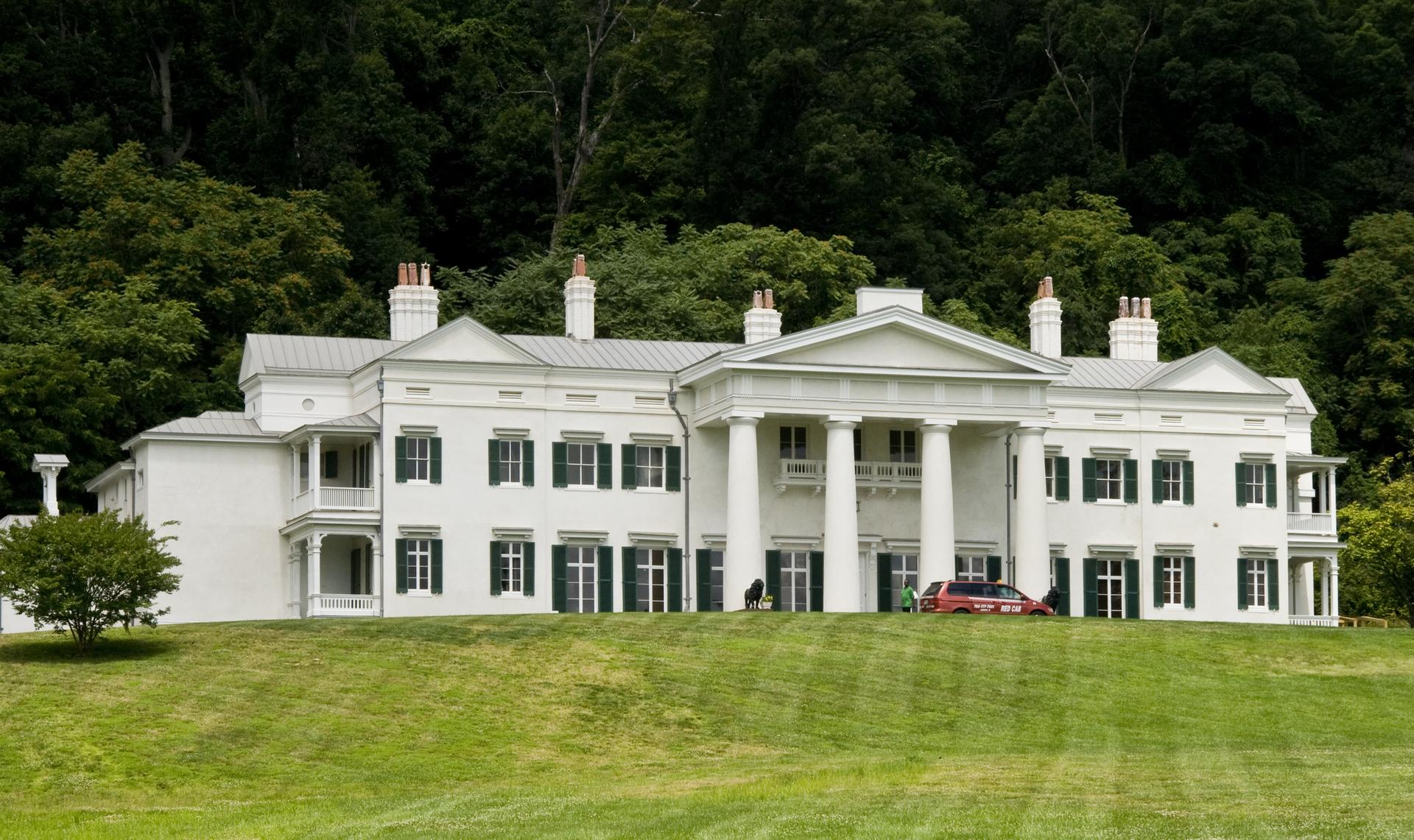 Large white mansion on a grassy hill, with a car and people, backed by forest.