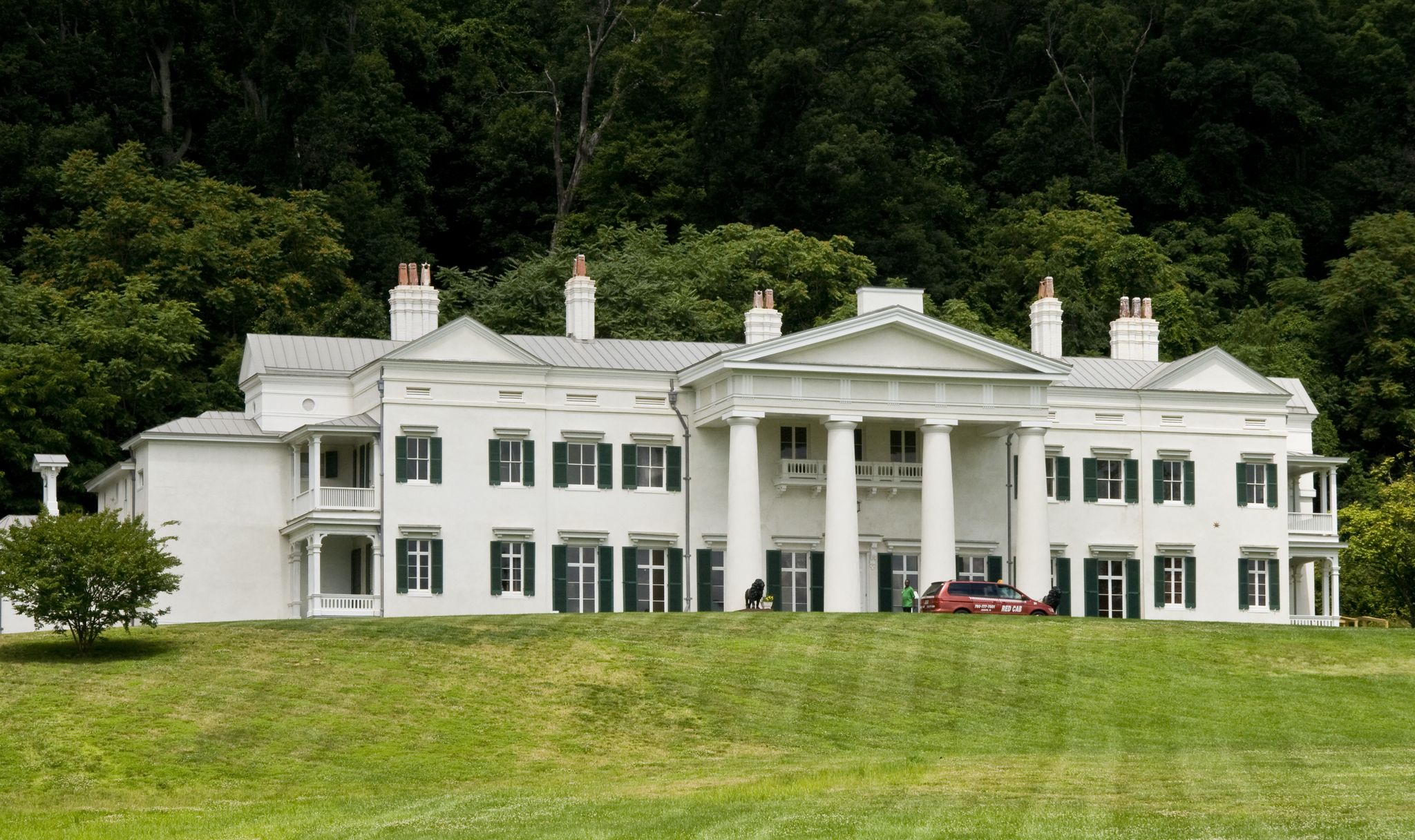 Large white mansion on a grassy hill, with a car and people, backed by forest. - Movan Park Loop