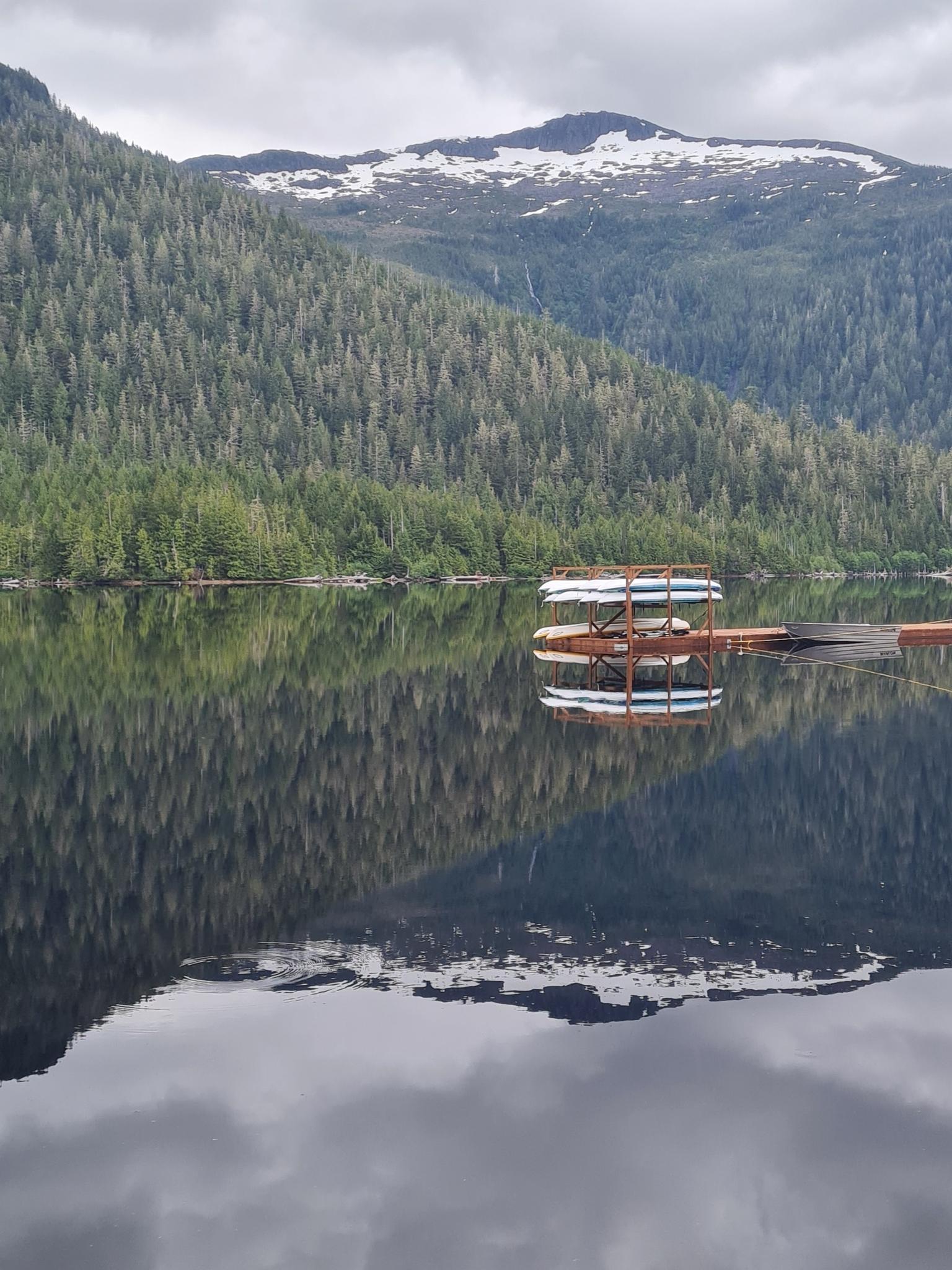 Calm lake reflecting forested mountains under an overcast sky, kayaks on a dock.
