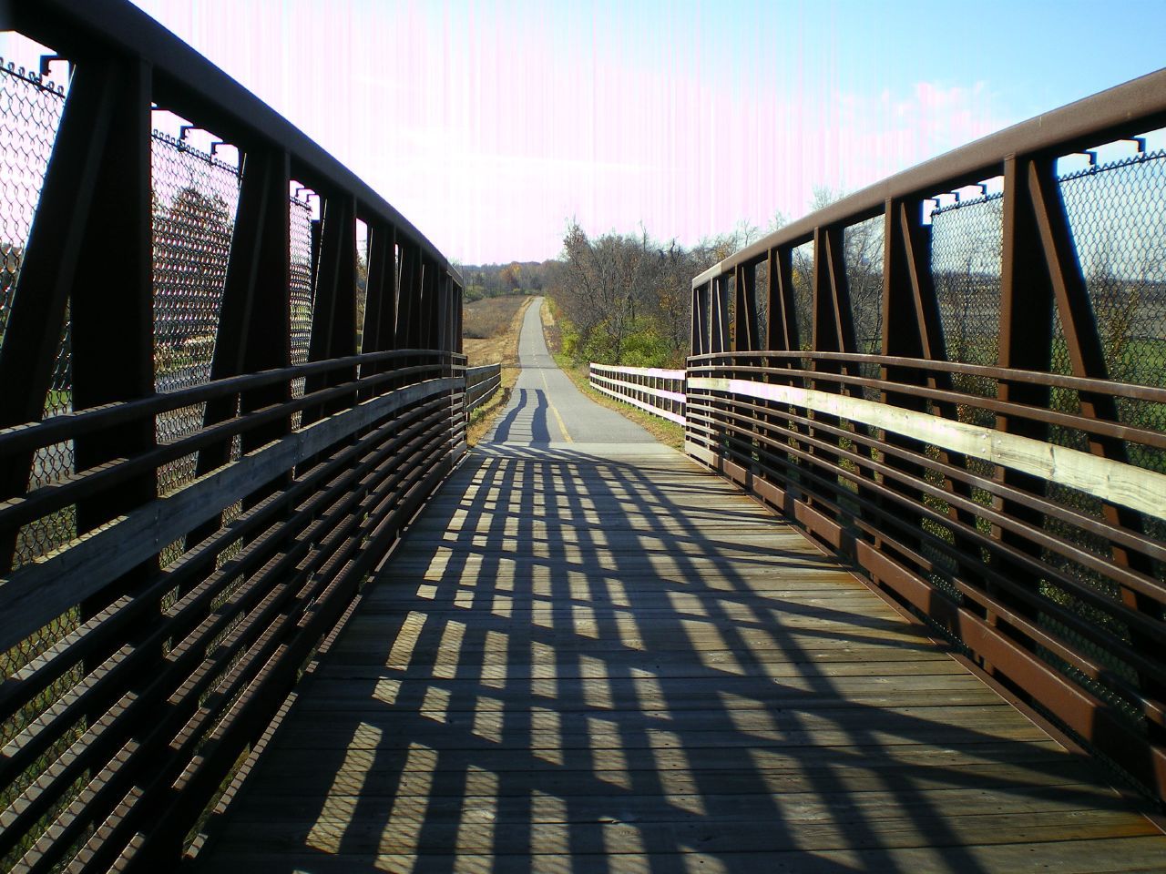Sunny view from a wooden bridge with strong shadows onto a paved road. - MCT Schoolhouse Trail