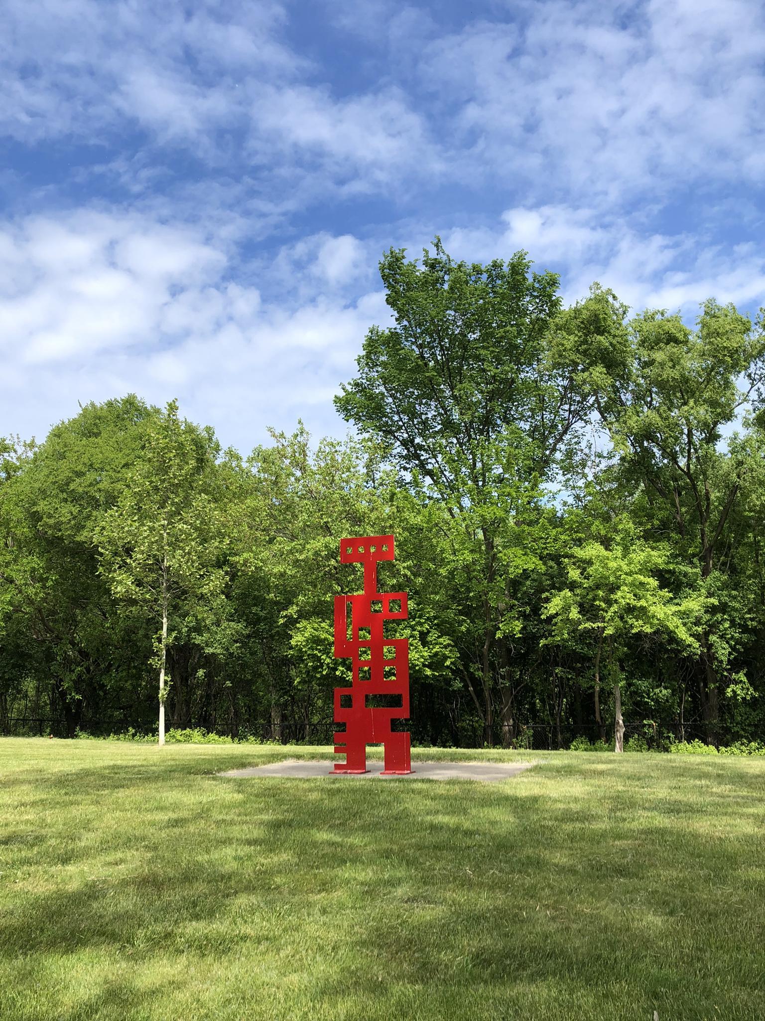 A tall, red abstract sculpture stands in a green field under a cloudy sky.