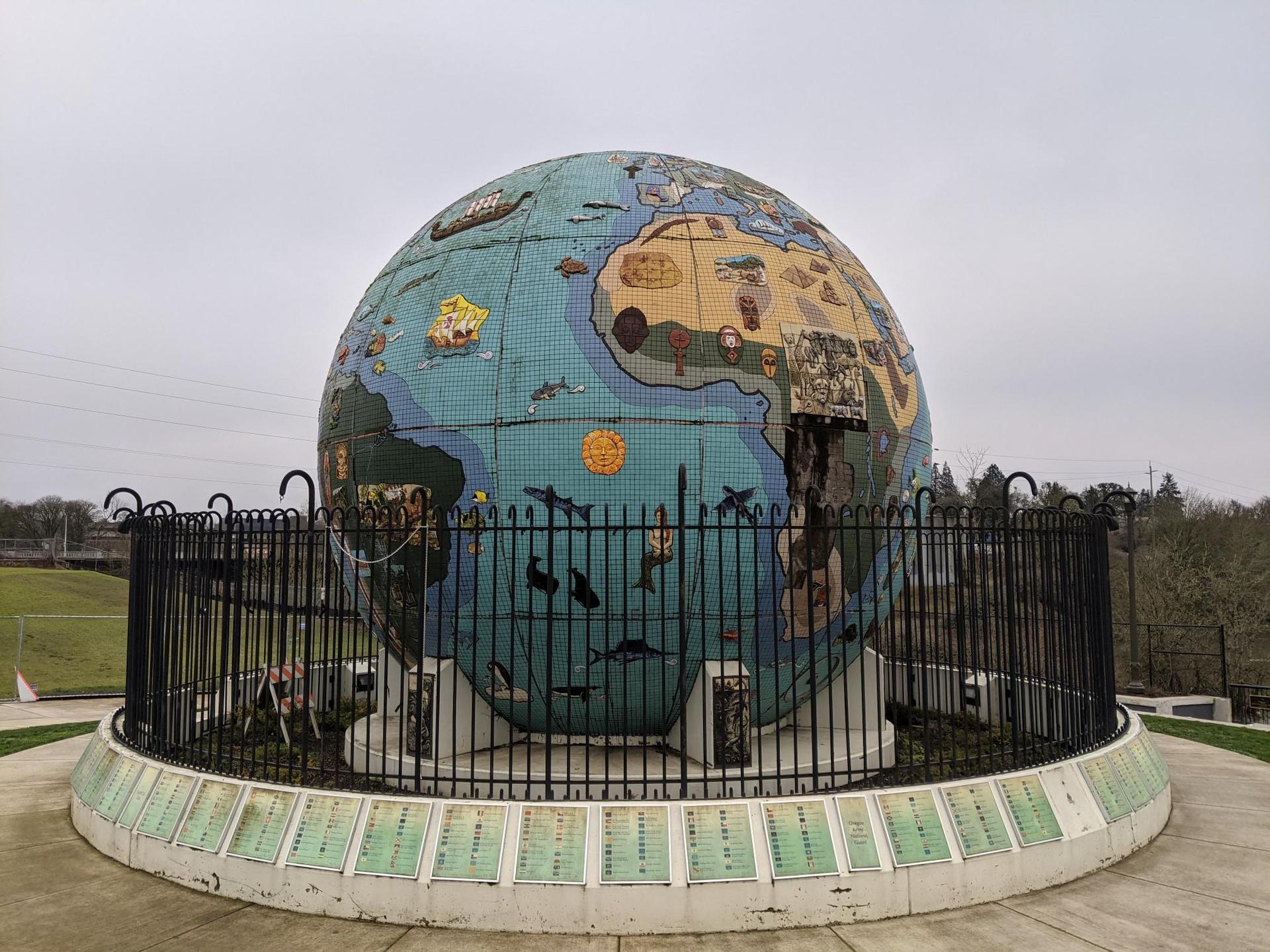 Large mosaic globe sculpture under an overcast sky, surrounded by a fence.