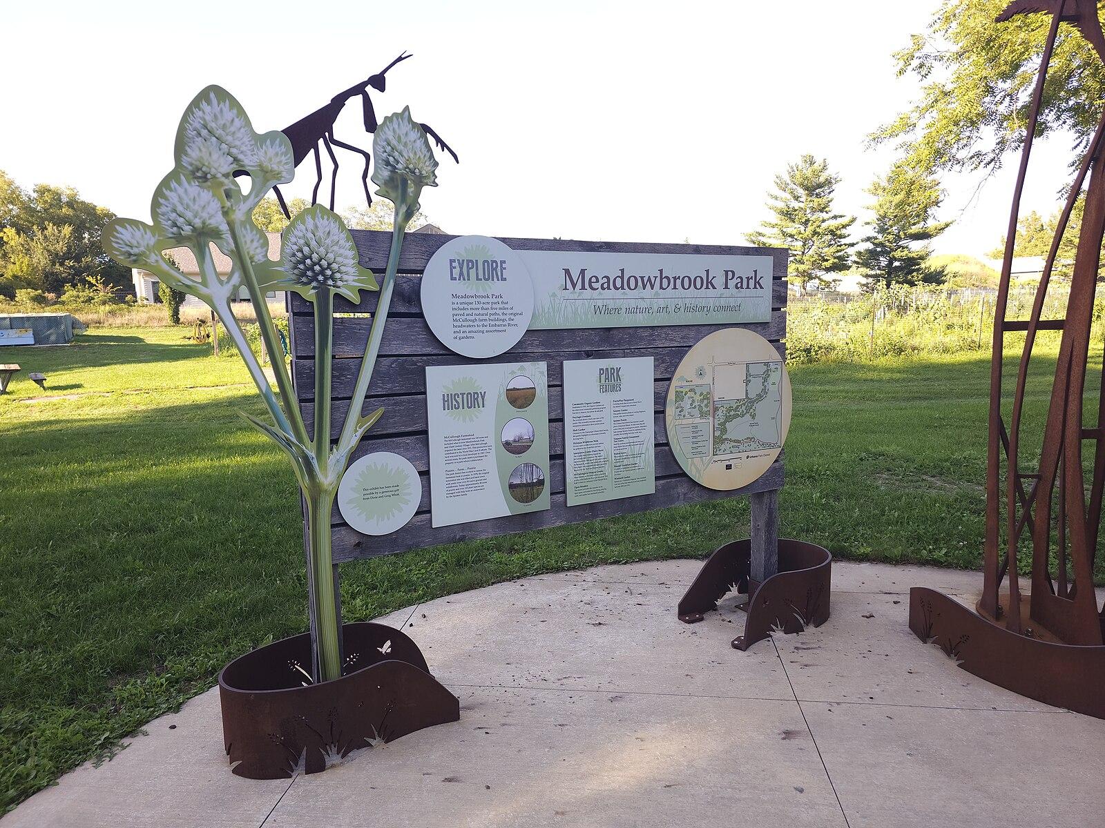 Outdoor information sign with metal plant and insect sculptures on a sunny day.