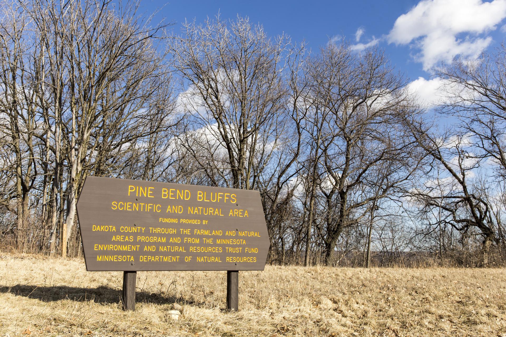Brown wooden sign in a dry field with bare trees under a blue sky.