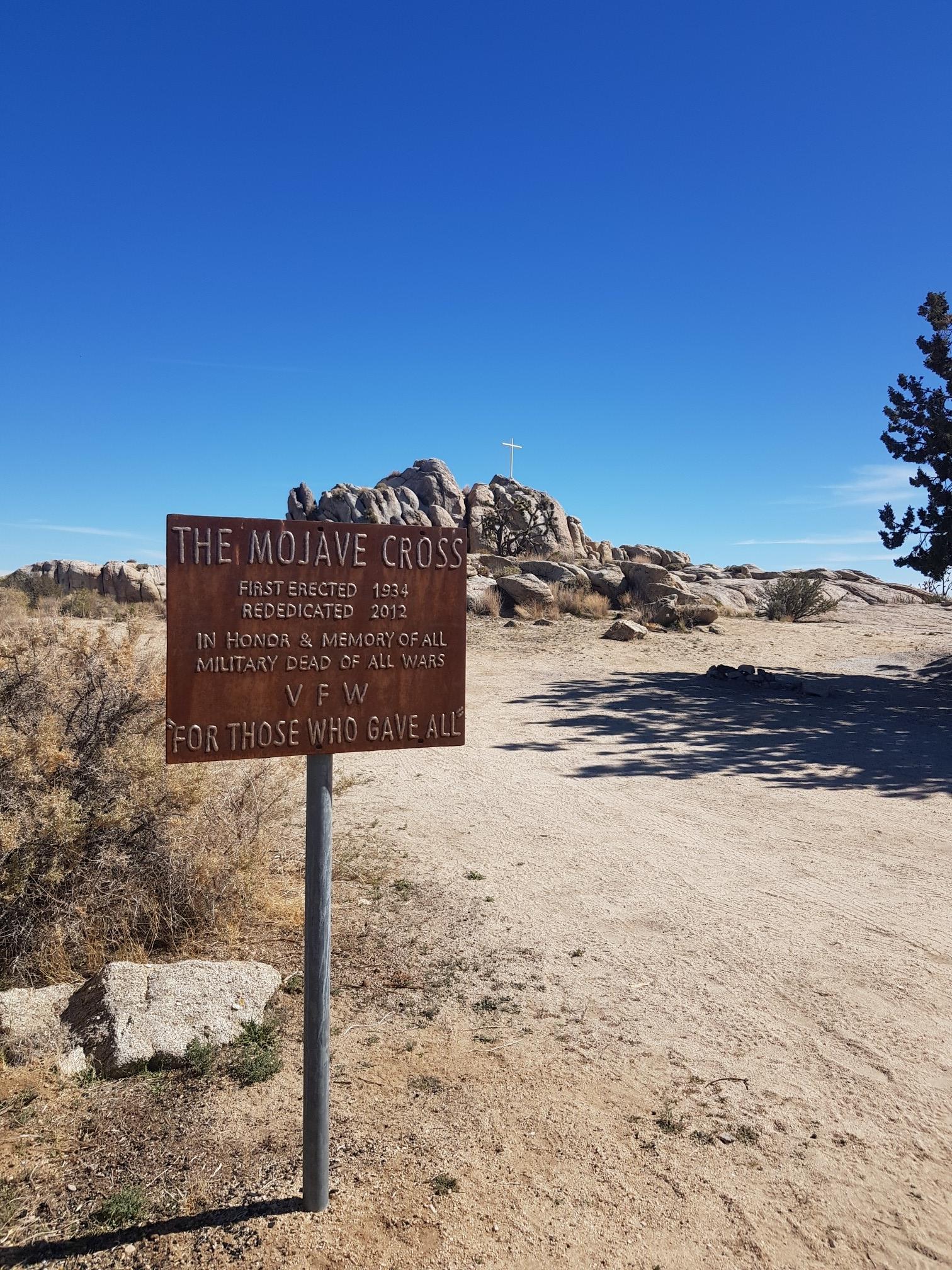 Metal sign on dirt path, distant cross on rocky hill.