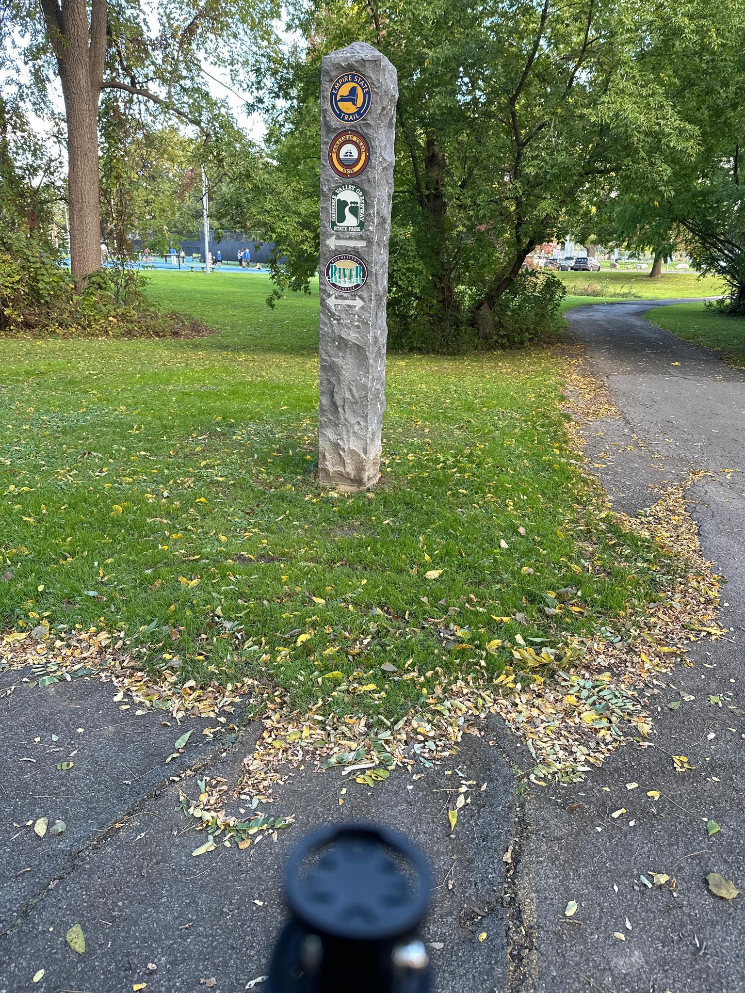 Stone marker with emblems beside a paved path, surrounded by grass and fallen leaves.