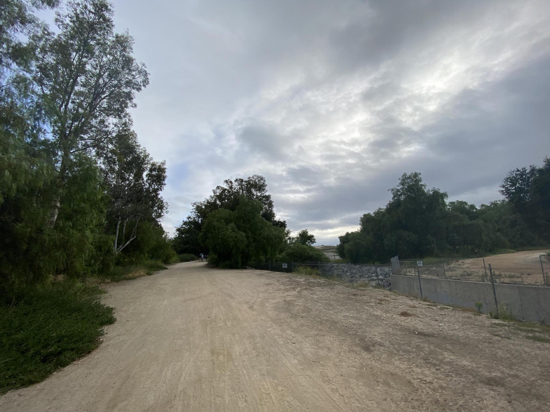 A wide dirt road under a cloudy sky, flanked by trees and a fence.